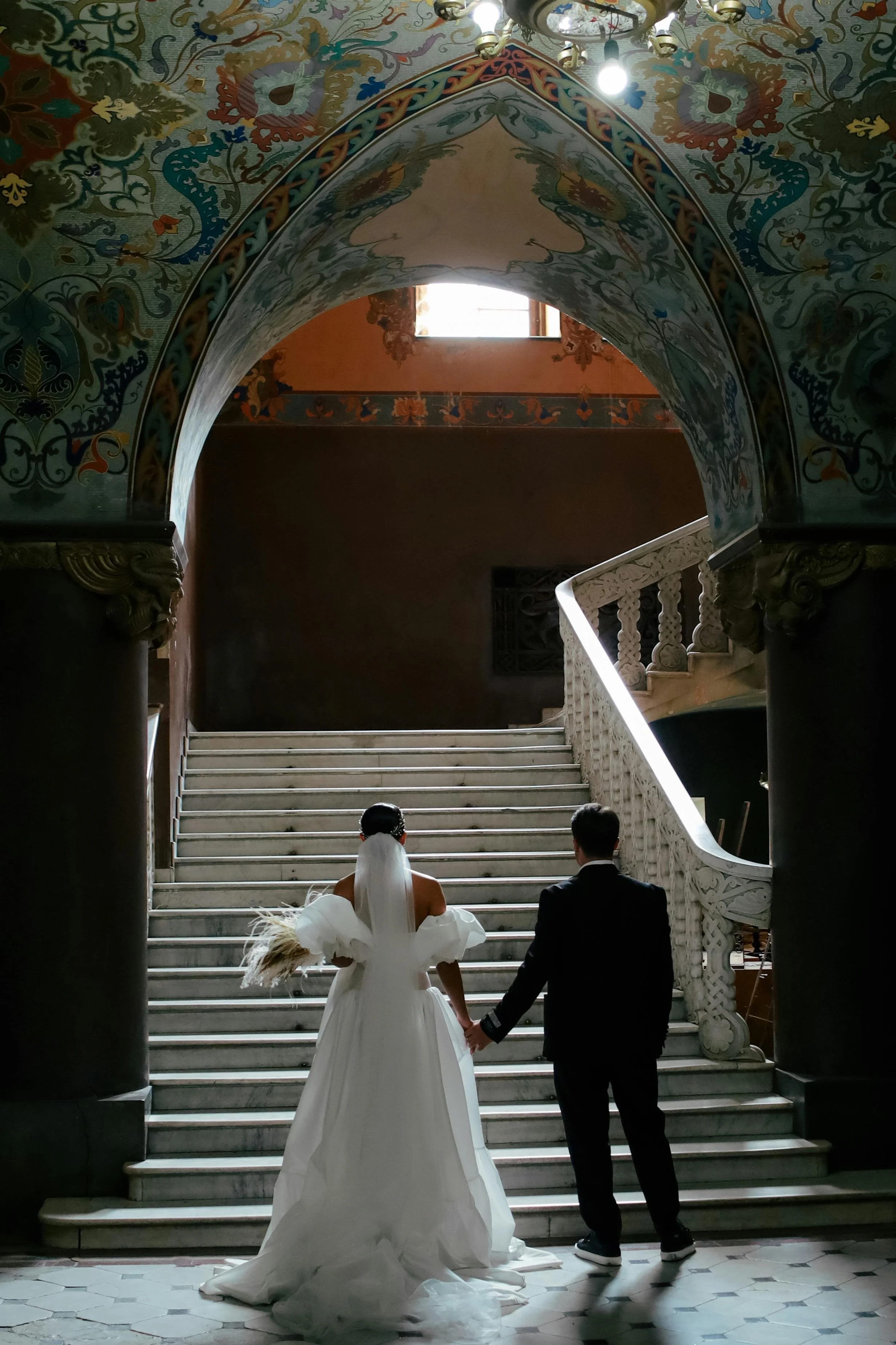 A bride and groom holding hands and walking stairs in a grand, ornately decorated building.