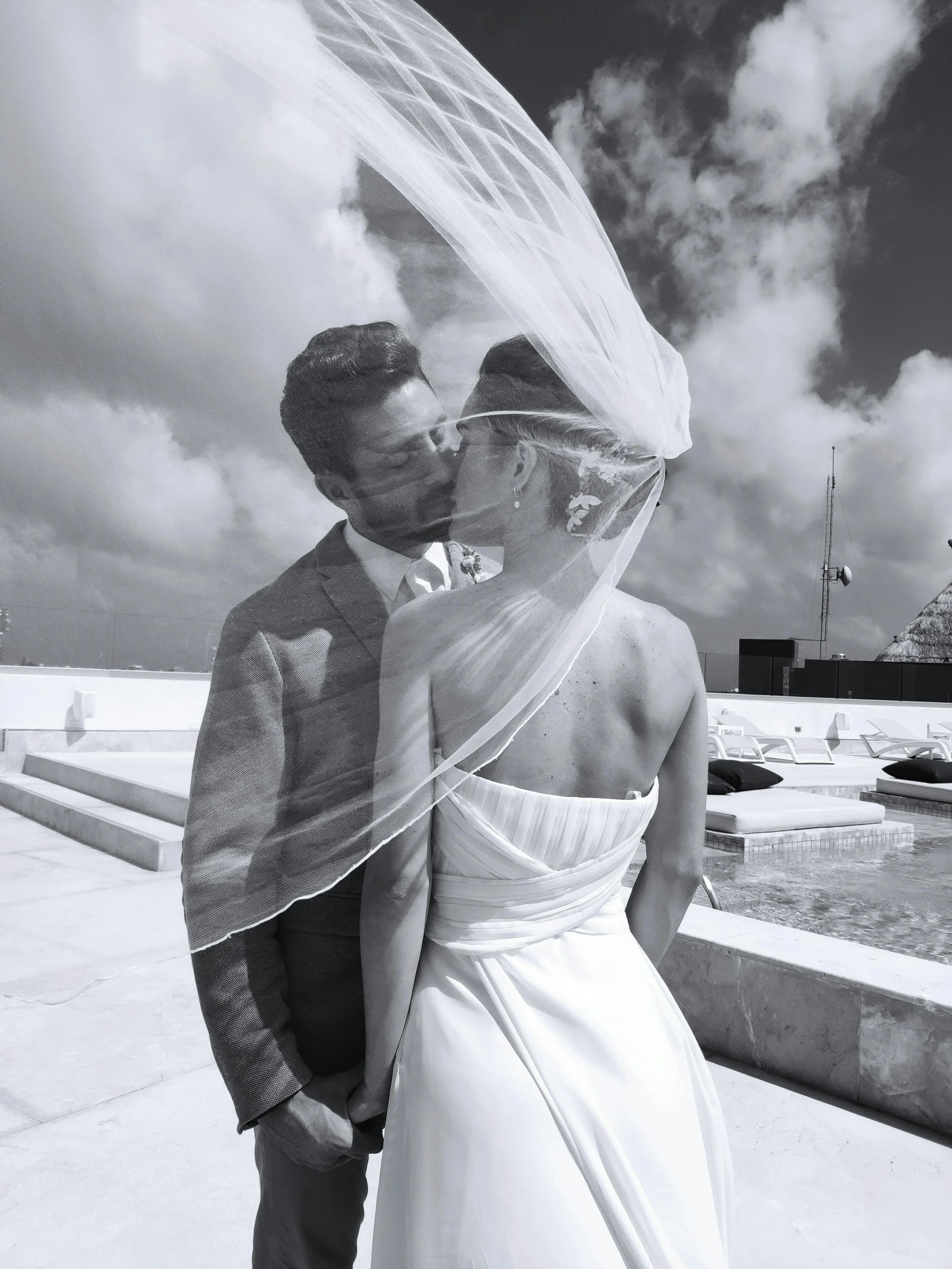 A black and white photo of a couple in wedding attire sharing a kiss on a rooftop with poolside lounge chairs and a cloudy sky in the background.