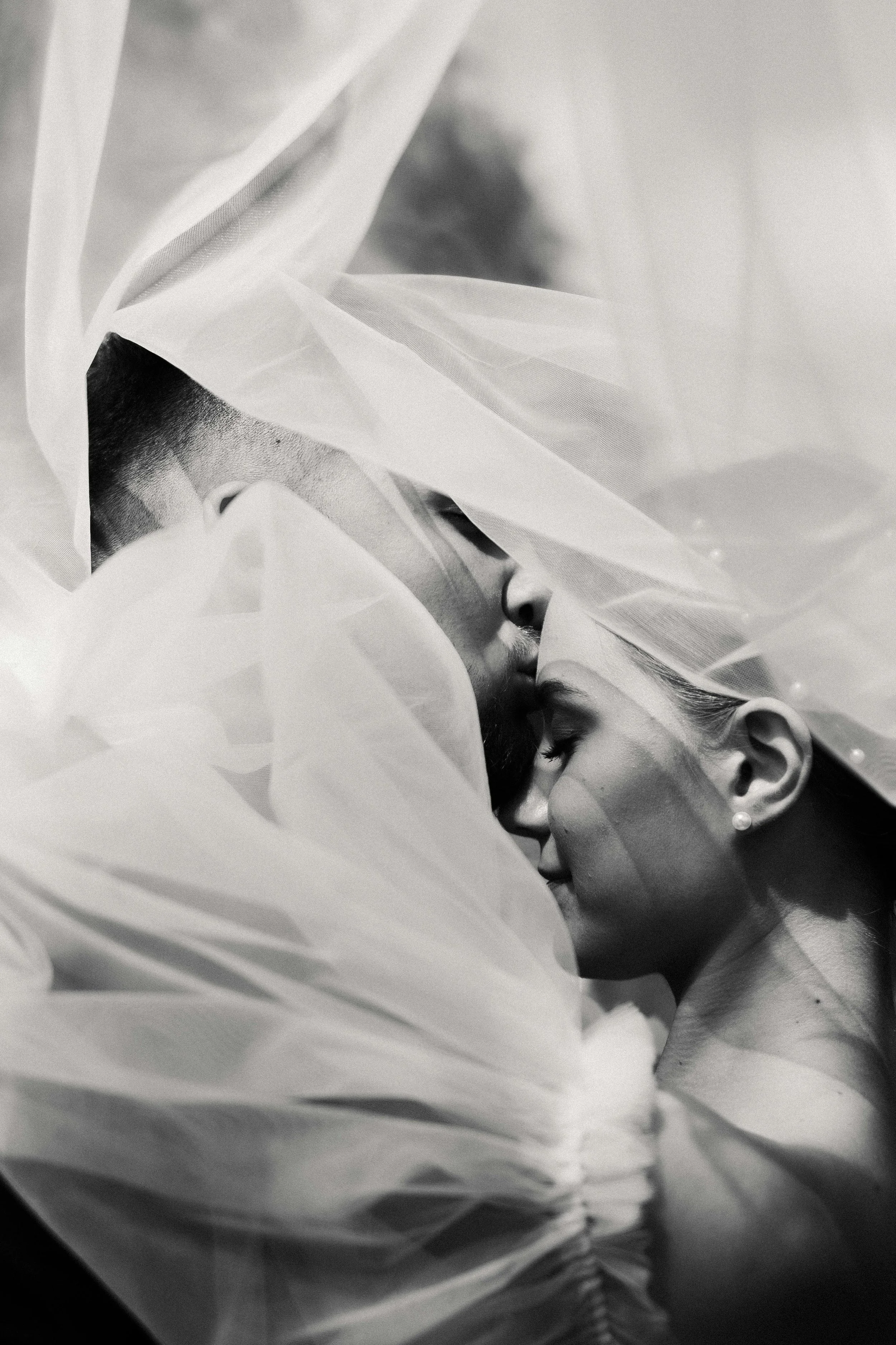 A black and white photograph of a romantic moment between a man and a woman, with their faces close together and eyes closed, surrounded by flowing sheer fabric.