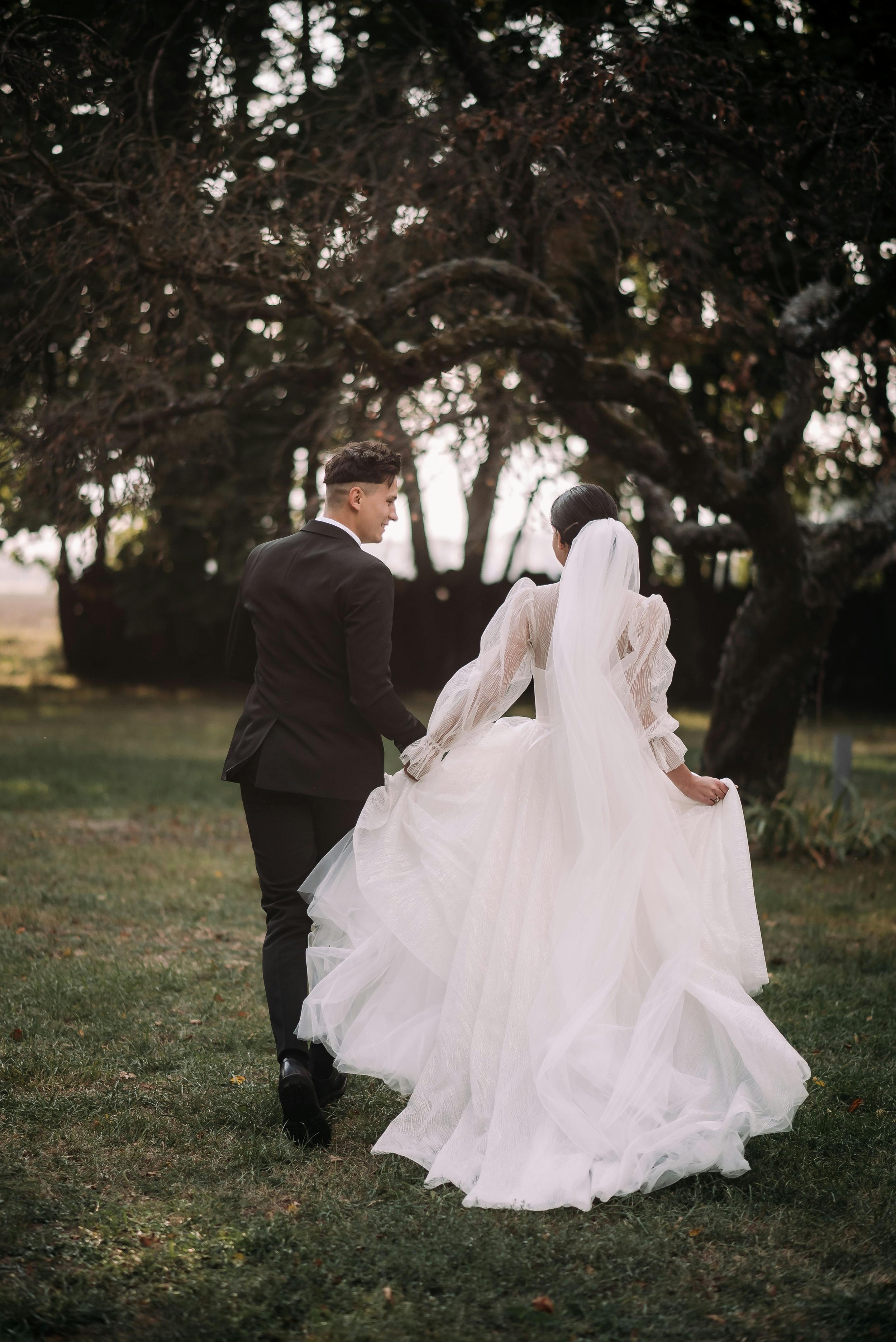 A bride and groom walking hand in hand outdoors, with the bride wearing a white wedding gown and veil, and the groom in a black suit, near a large tree with dark leaves.