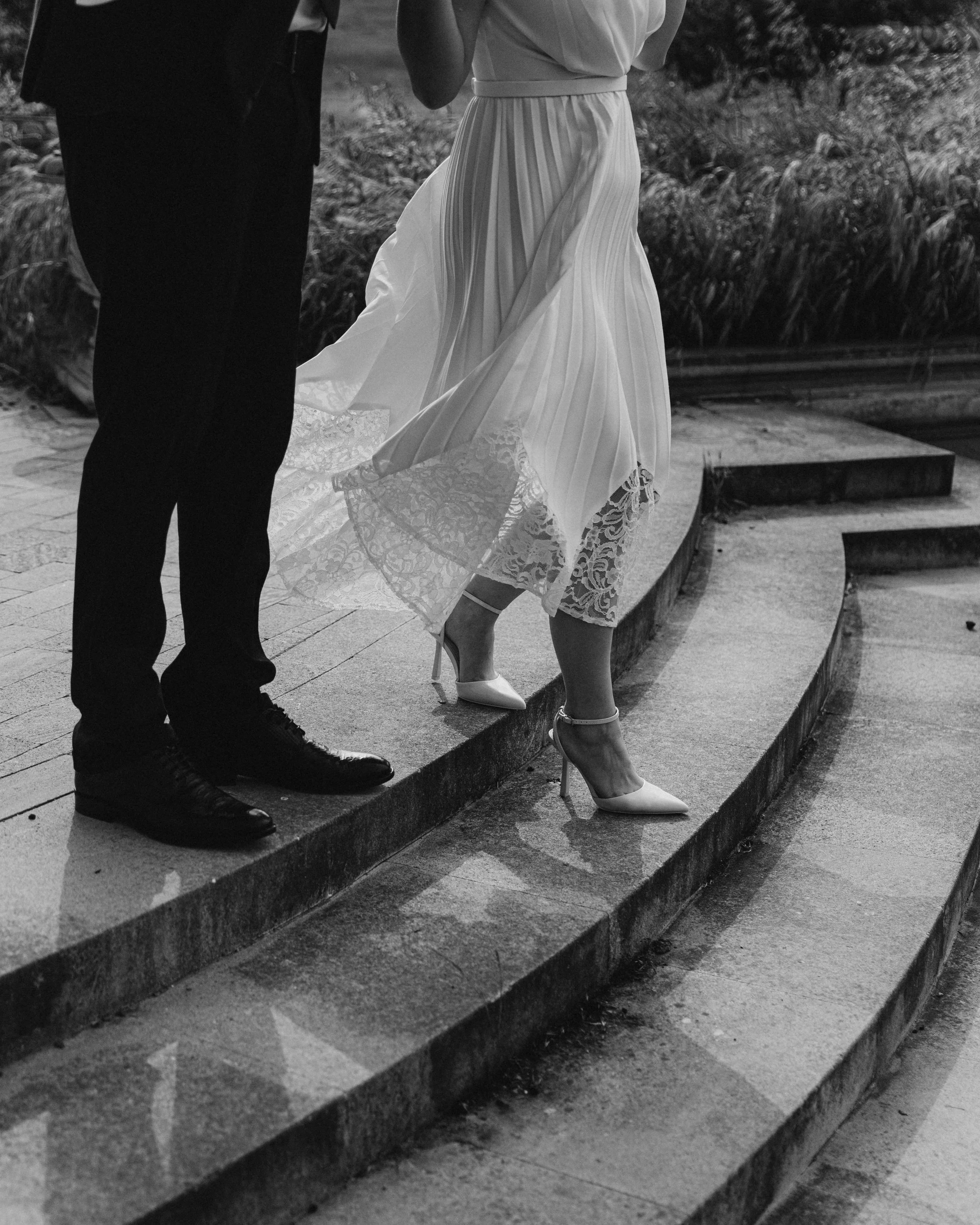 Close-up of a bride in a wedding dress and high heels, standing on steps next to a man in a suit, outdoors