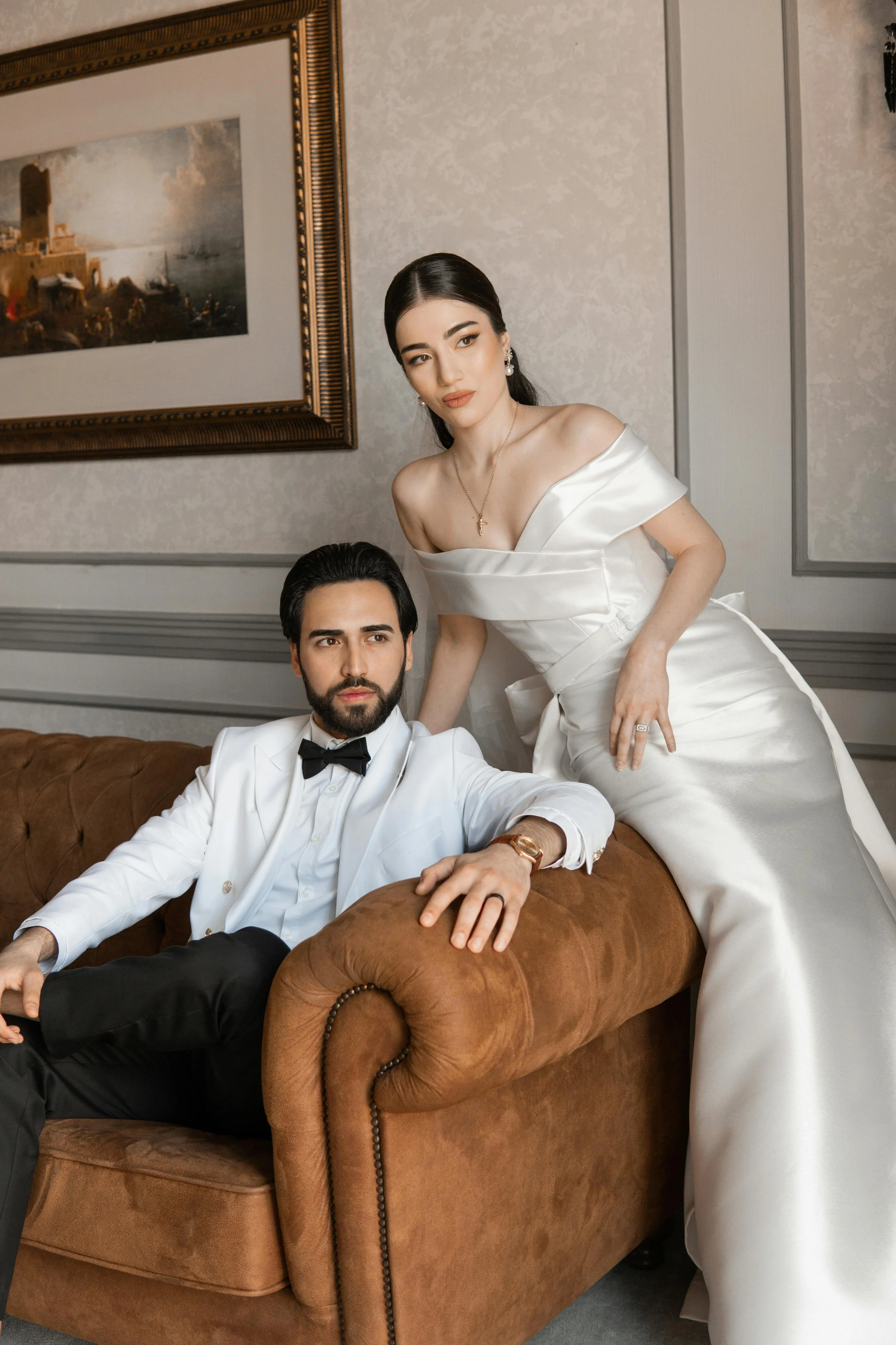 A man in a tuxedo with a bow tie and a woman in a white satin wedding dress sitting and standing on a vintage brown sofa in an elegant room with framed artwork on the wall.