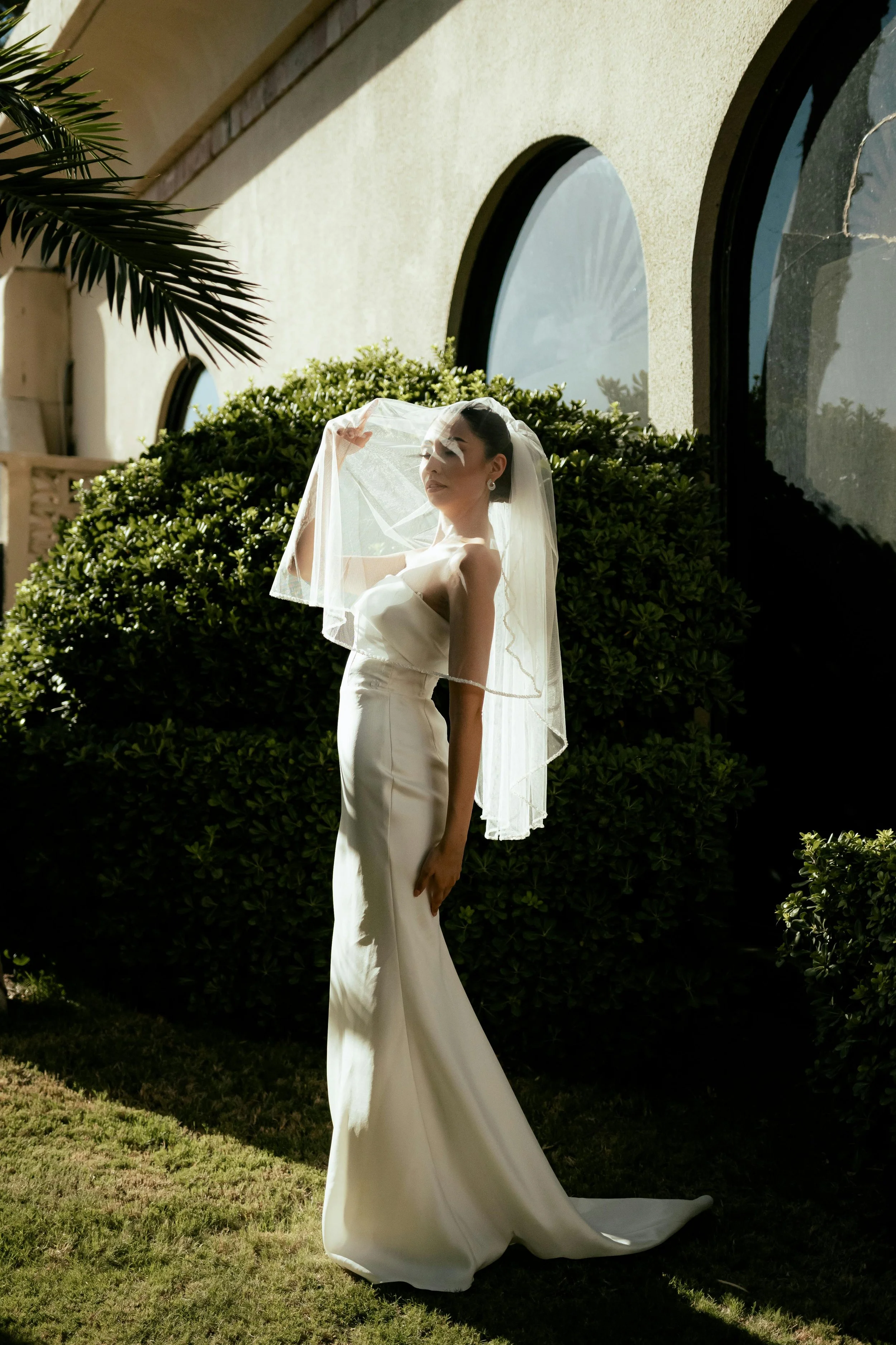 A bride in a strapless white wedding gown stands outdoors near green bushes, holding a sheer veil over her face with sunlight highlighting her profile.