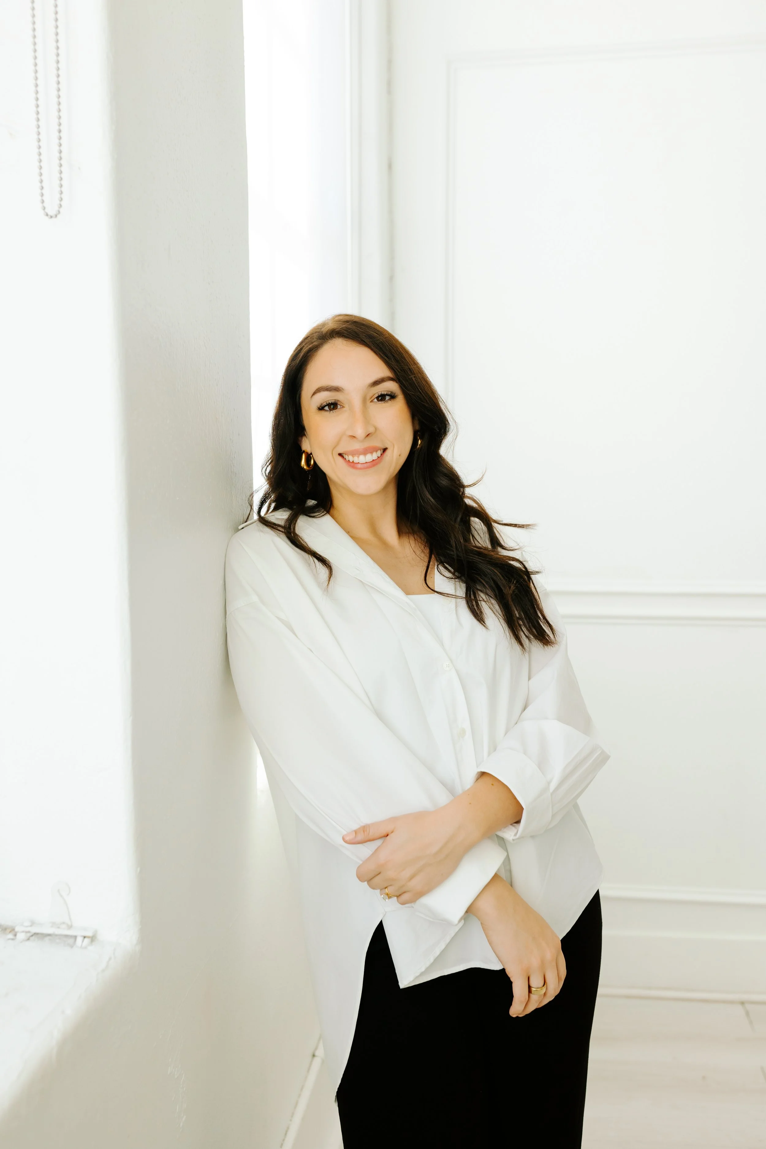 A woman with long, dark hair and a bright smile, wearing a white shirt and black pants, leaning against a white wall in a brightly lit indoor space.