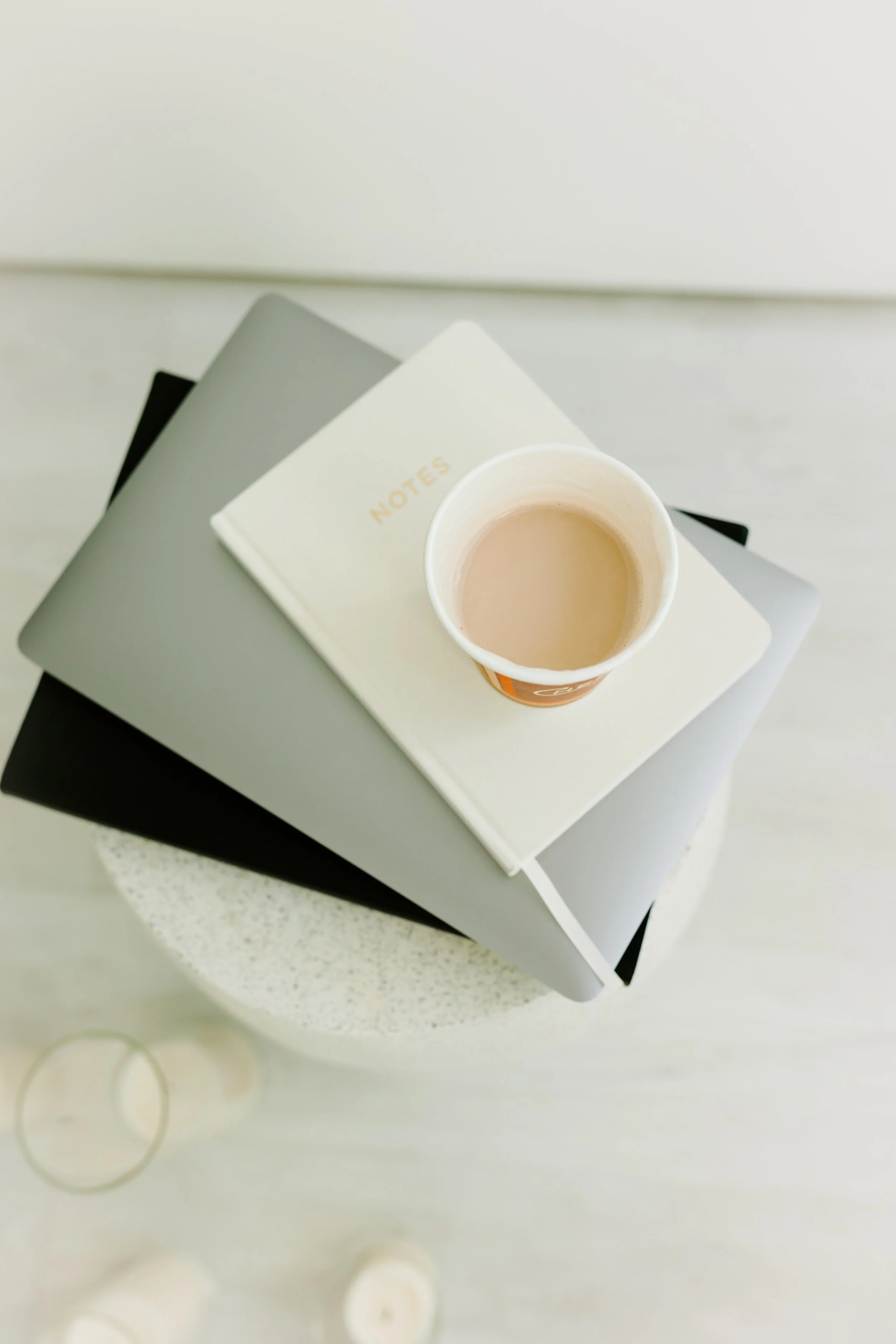 A top-down view of a white coffee cup with a hot beverage, placed on a stack of notebooks and magazines on a small round table.