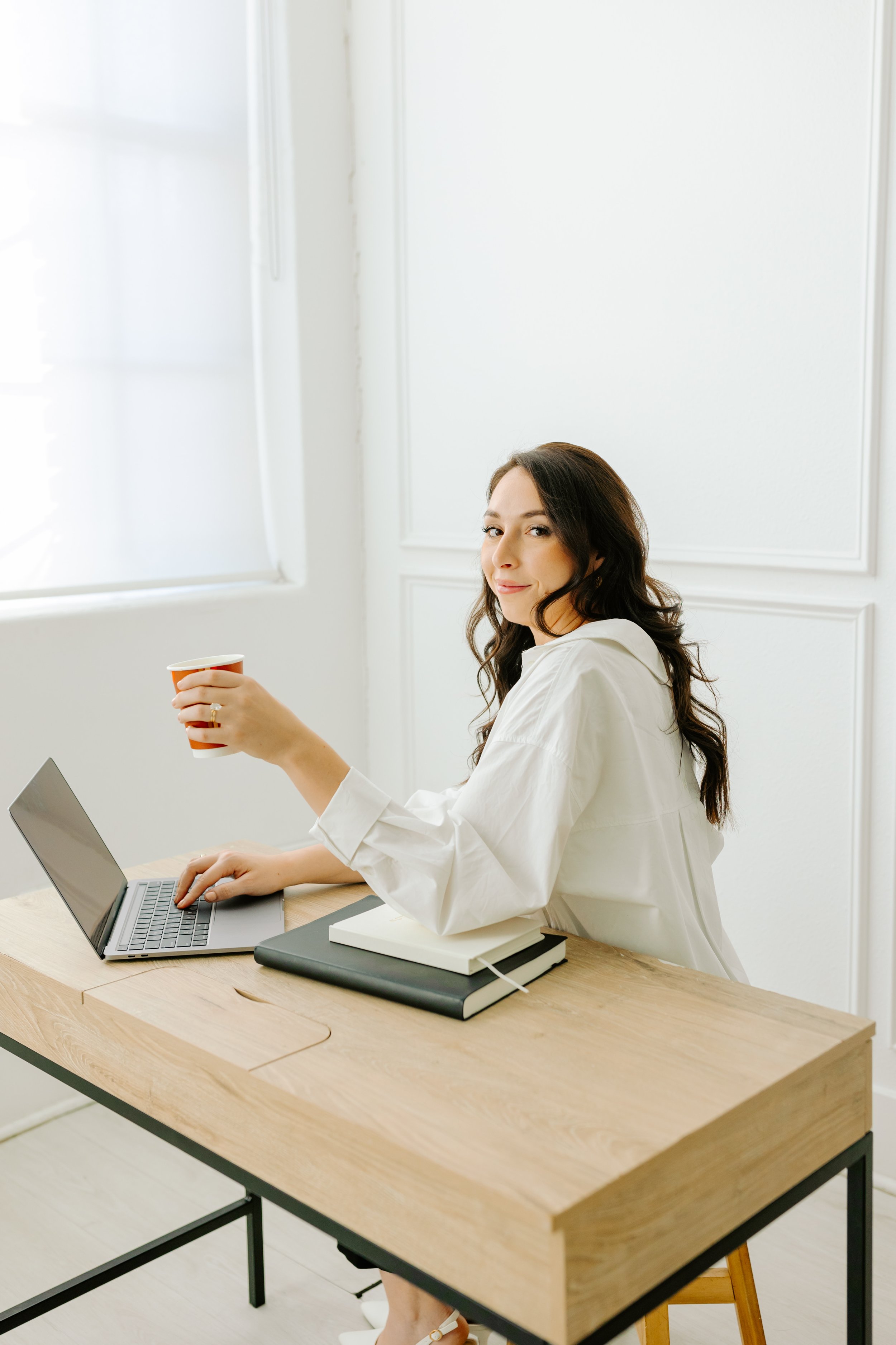 Woman sitting at a desk with a laptop, holding a cup, in a bright modern room.