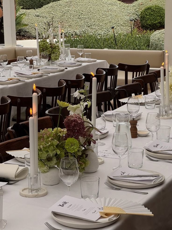 A beautifully set dining table with white tablecloth, floral centerpiece, candles, and glassware, outdoors with a garden view in the background.