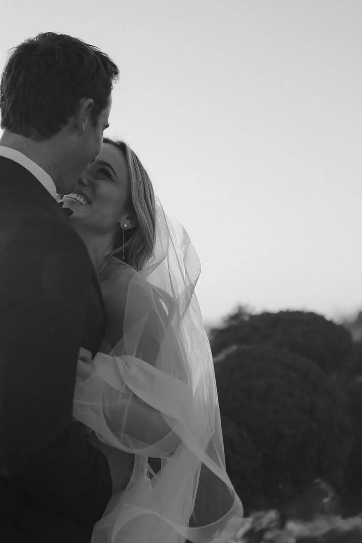 A black and white photo of a bride and groom smiling and looking at each other outdoors, with trees in the background.