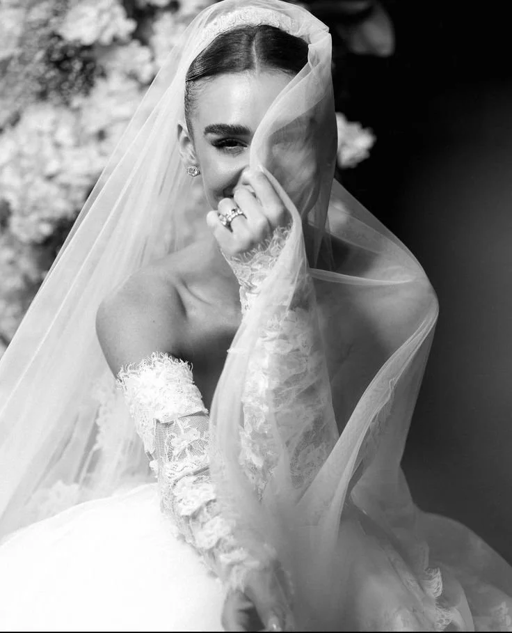 A bride in a wedding dress and veil covering half her face, smiling and holding the veil with her hand, wearing rings and lace gloves, with a blurred background.