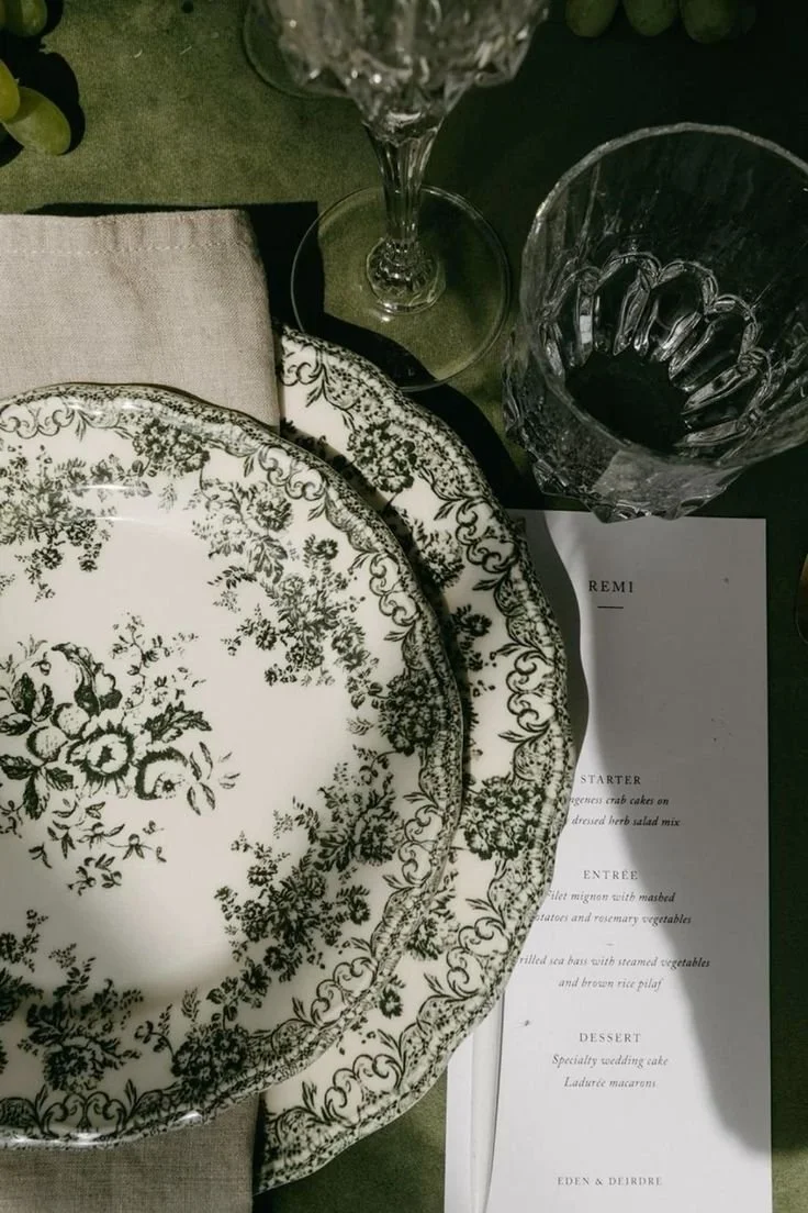 Black and white floral patterned dinnerware set including a large plate and a salad plate on a table with a menu, a cloth napkin, empty wine glass, and a water glass.