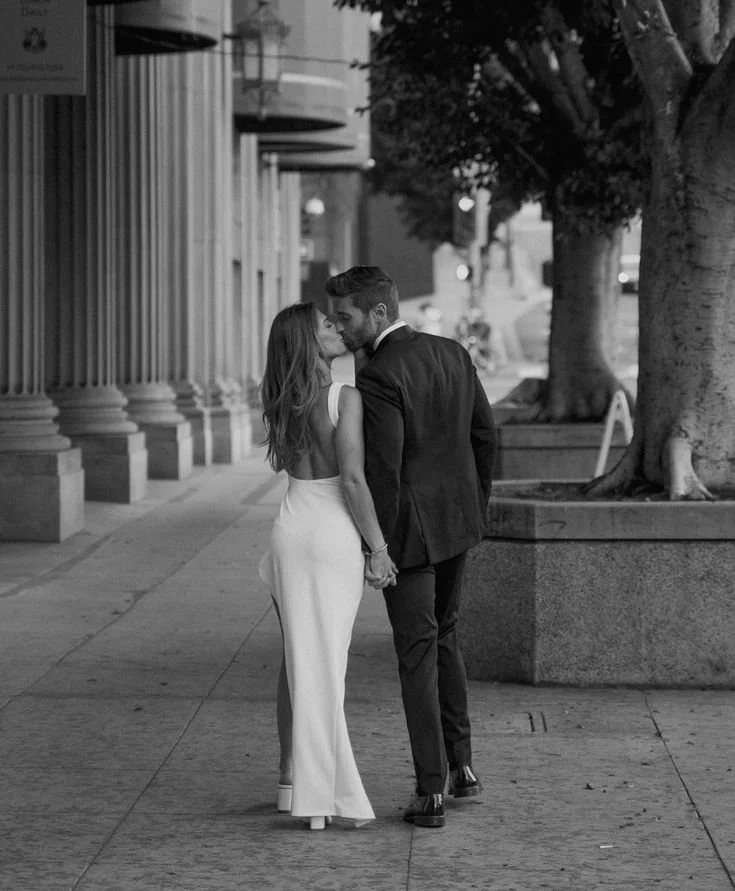 A couple dressed in formal attire holding hands and kissing on an urban sidewalk with trees and buildings in the background.