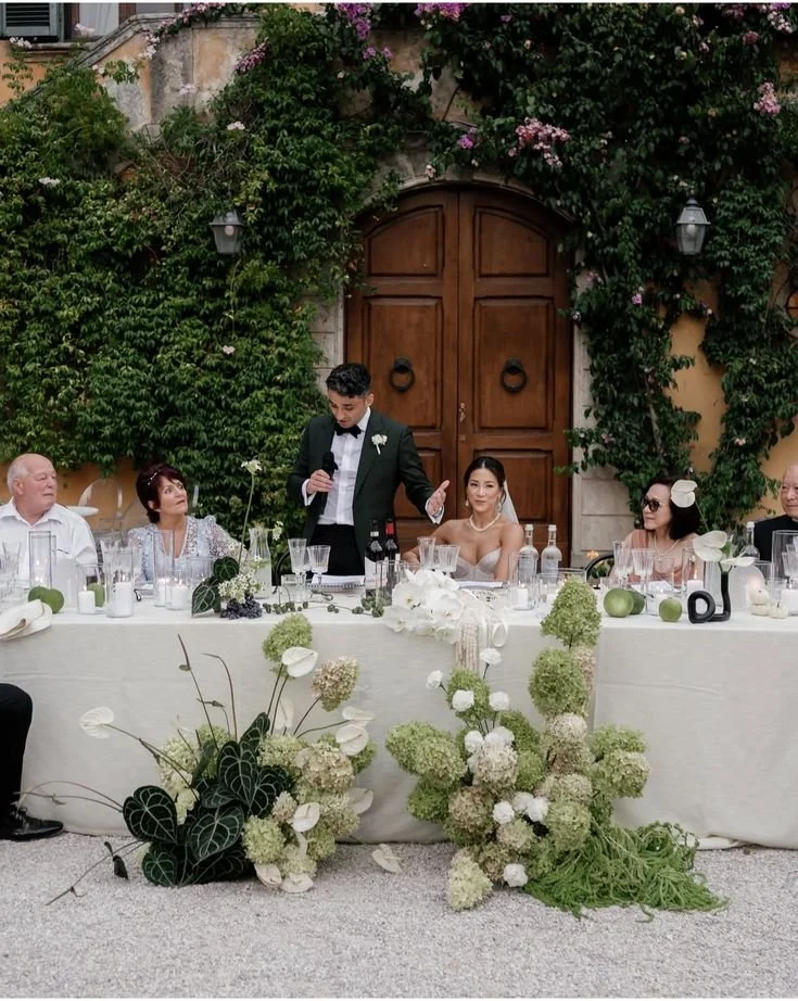 Groom giving a speech at a wedding reception, seated at a decorated table with bridesmaids and family, outdoors with a large wooden door and flower-covered walls in the background.