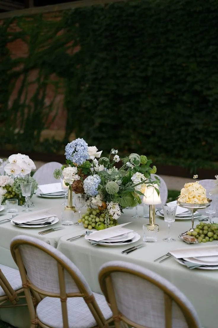 A decorated outdoor banquet table with white tablecloth, floral centerpiece, plates, silverware, glasses, and candles, set against a green hedge wall.