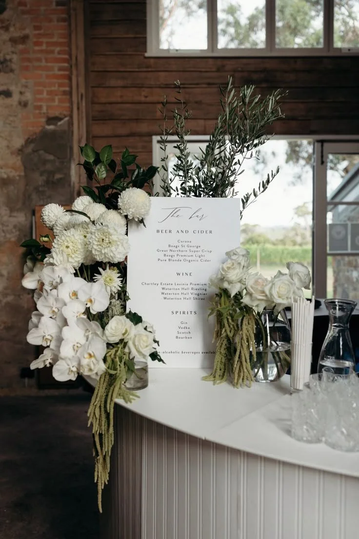 A wedding bar menu is decorated with white flowers and green foliage on a white table, with a rustic brick and wood interior in the background.