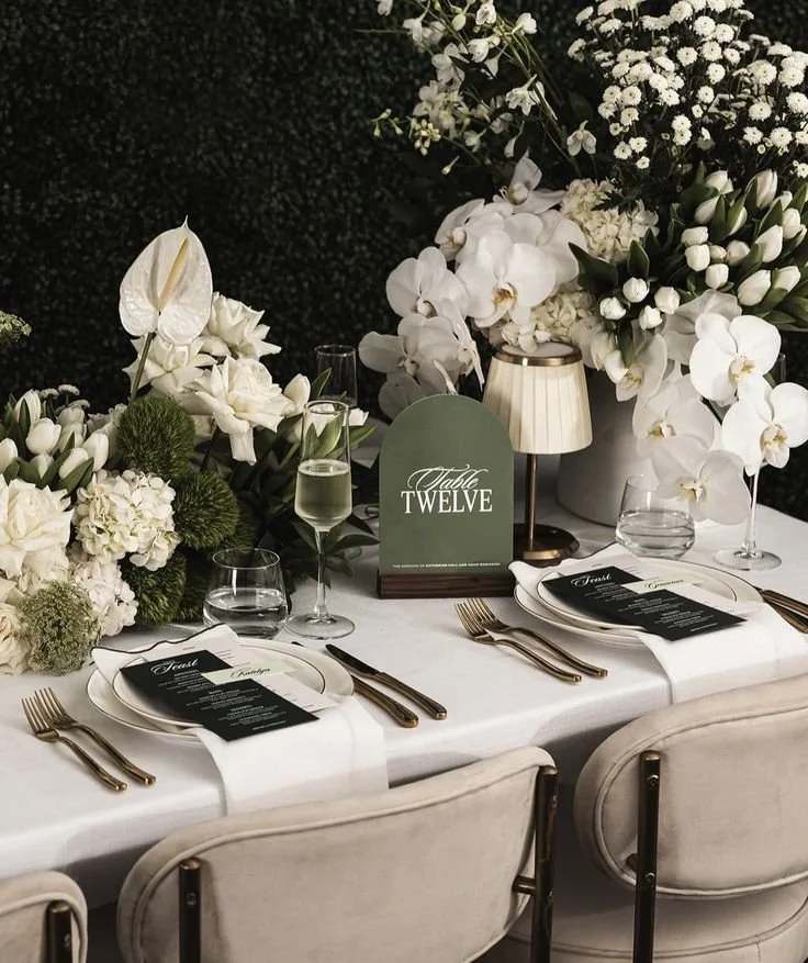 Elegant dining table with white tablecloth, floral arrangements, place settings, and a sign reading "Table Twelve."