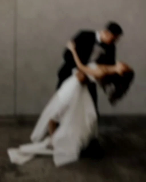 A couple dancing closely, the man in a suit and the woman in a white dress, against a gray background.
