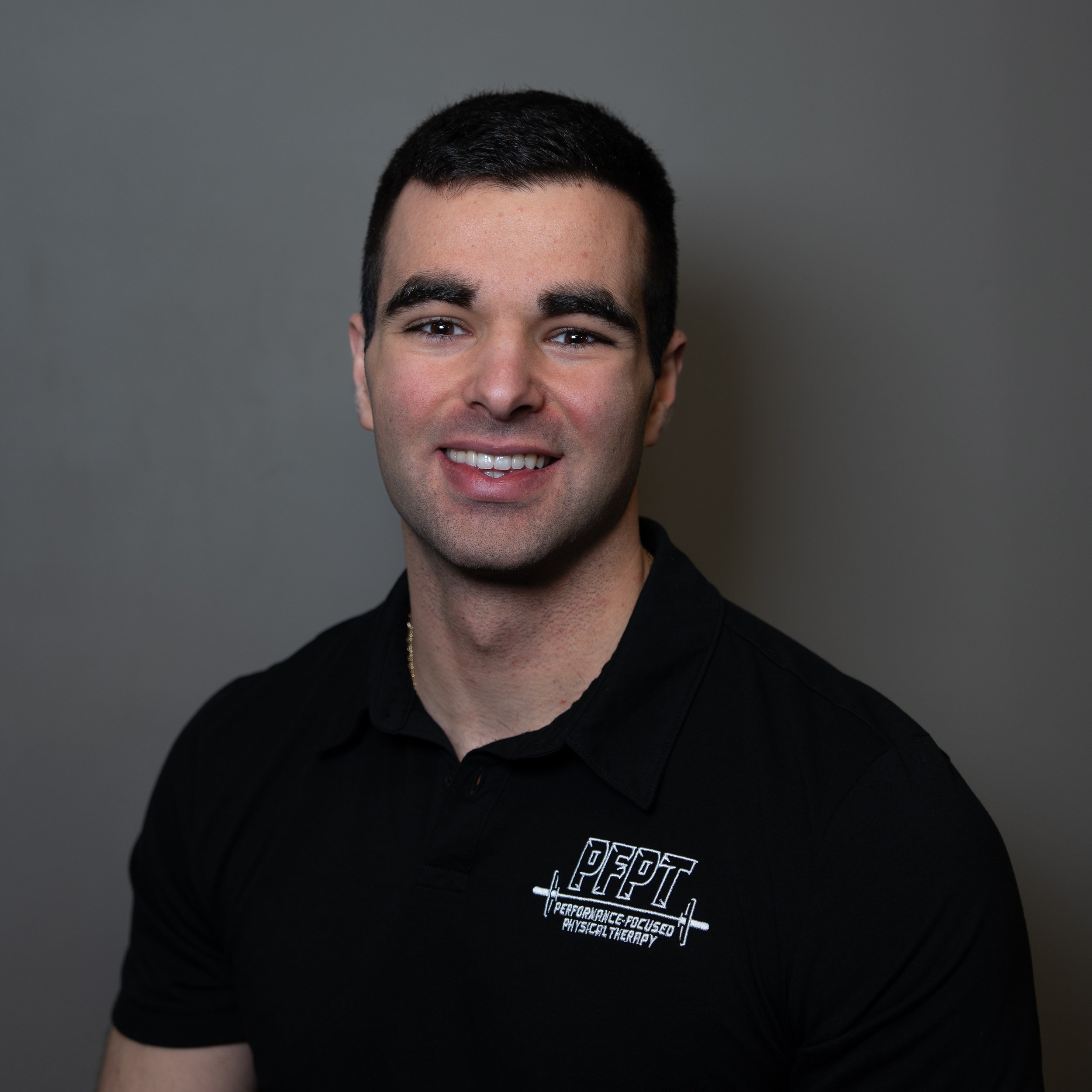 A young man with dark hair, smiling, wearing a black shirt with the logo 'PPCT' and the text 'Performance Focused Physical Therapy' on it, standing against a plain gray background.