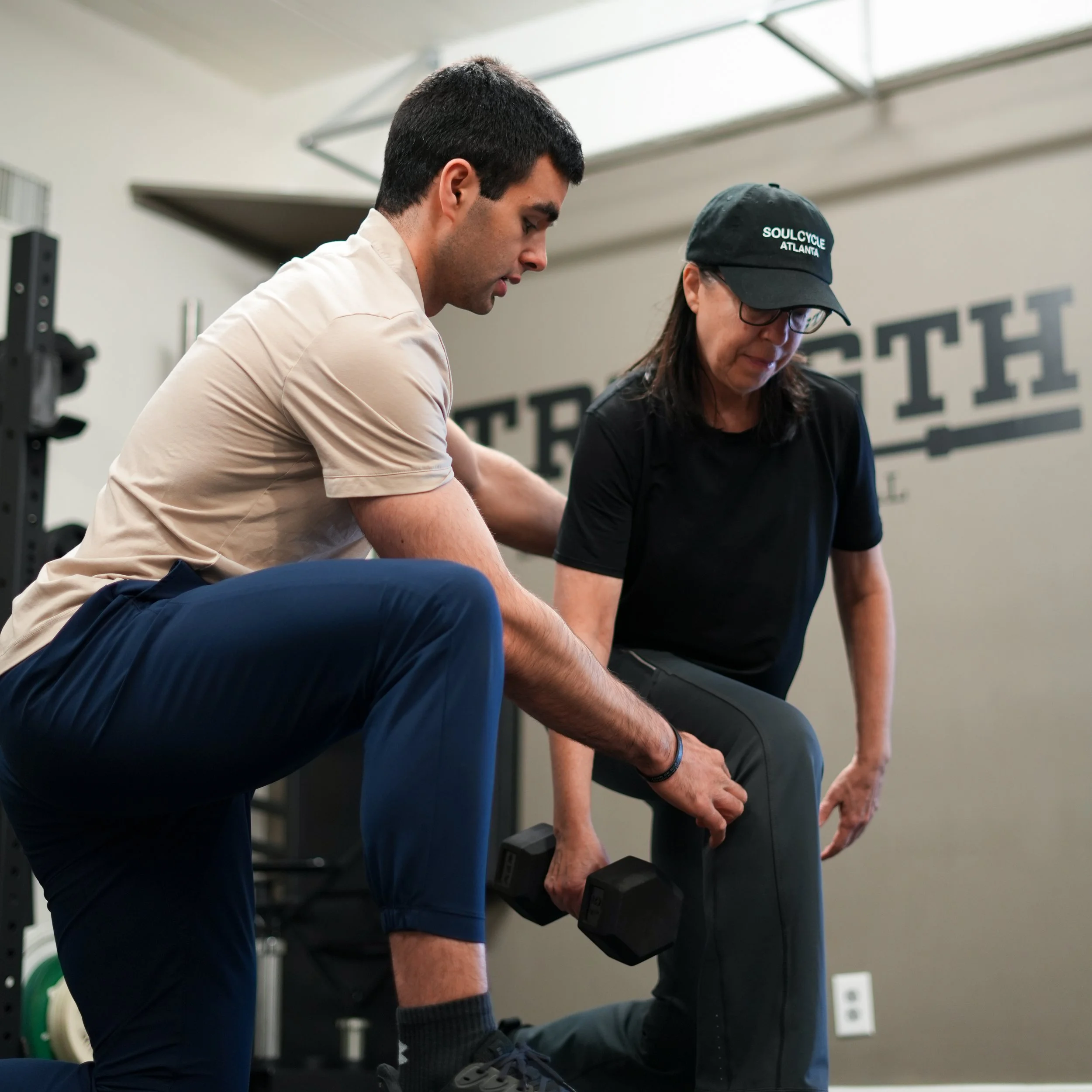 Two men are lifting dumbbells during a workout in a gym. One man is wearing a beige shirt and dark pants, and the other man is wearing a black shirt, black cap, and glasses. They are standing in front of a wall with horizontal lines.