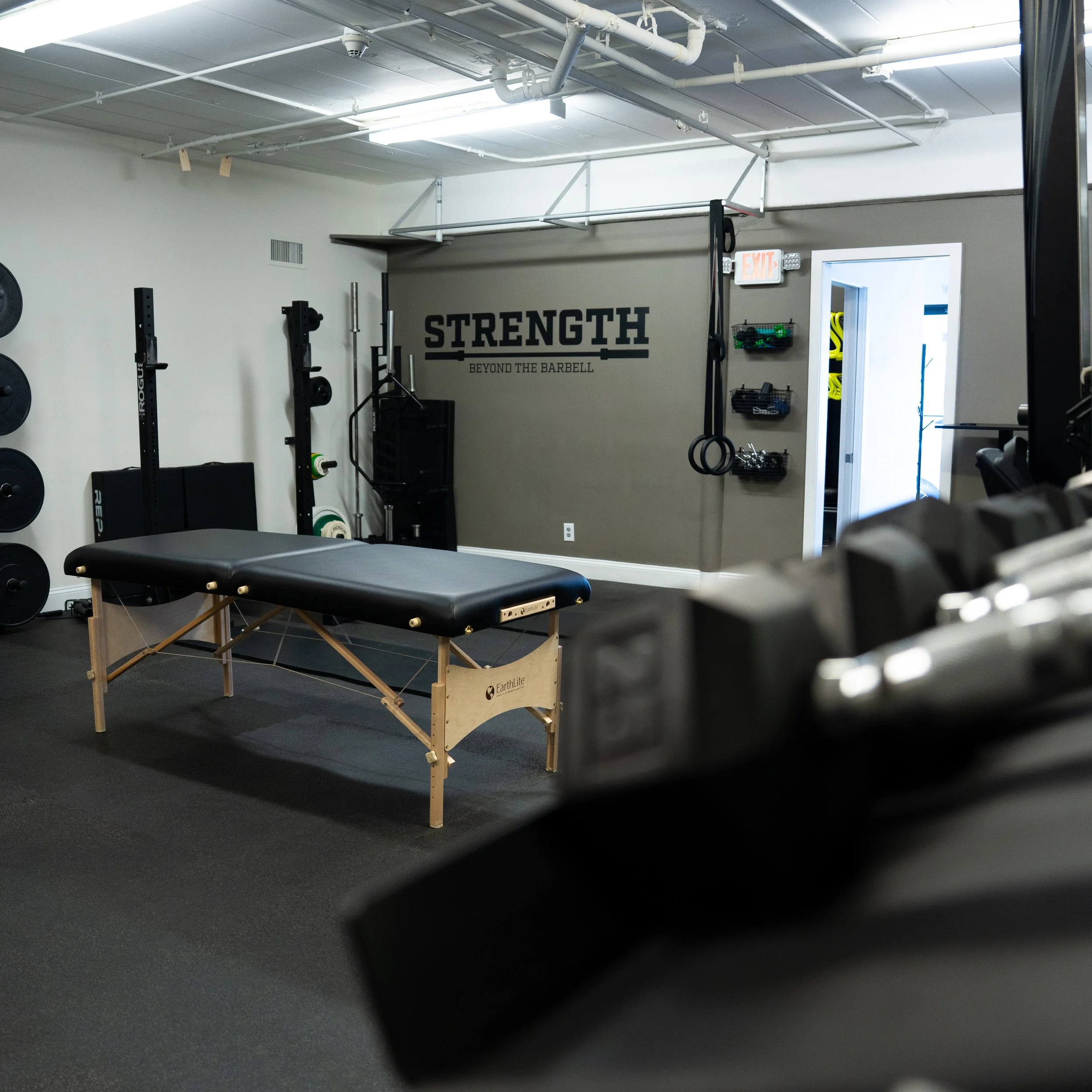 Interior of a gym with strength training equipment and a massage table, with a sign on the wall that reads 'STRENGTH BEYOND THE BARBELL'.