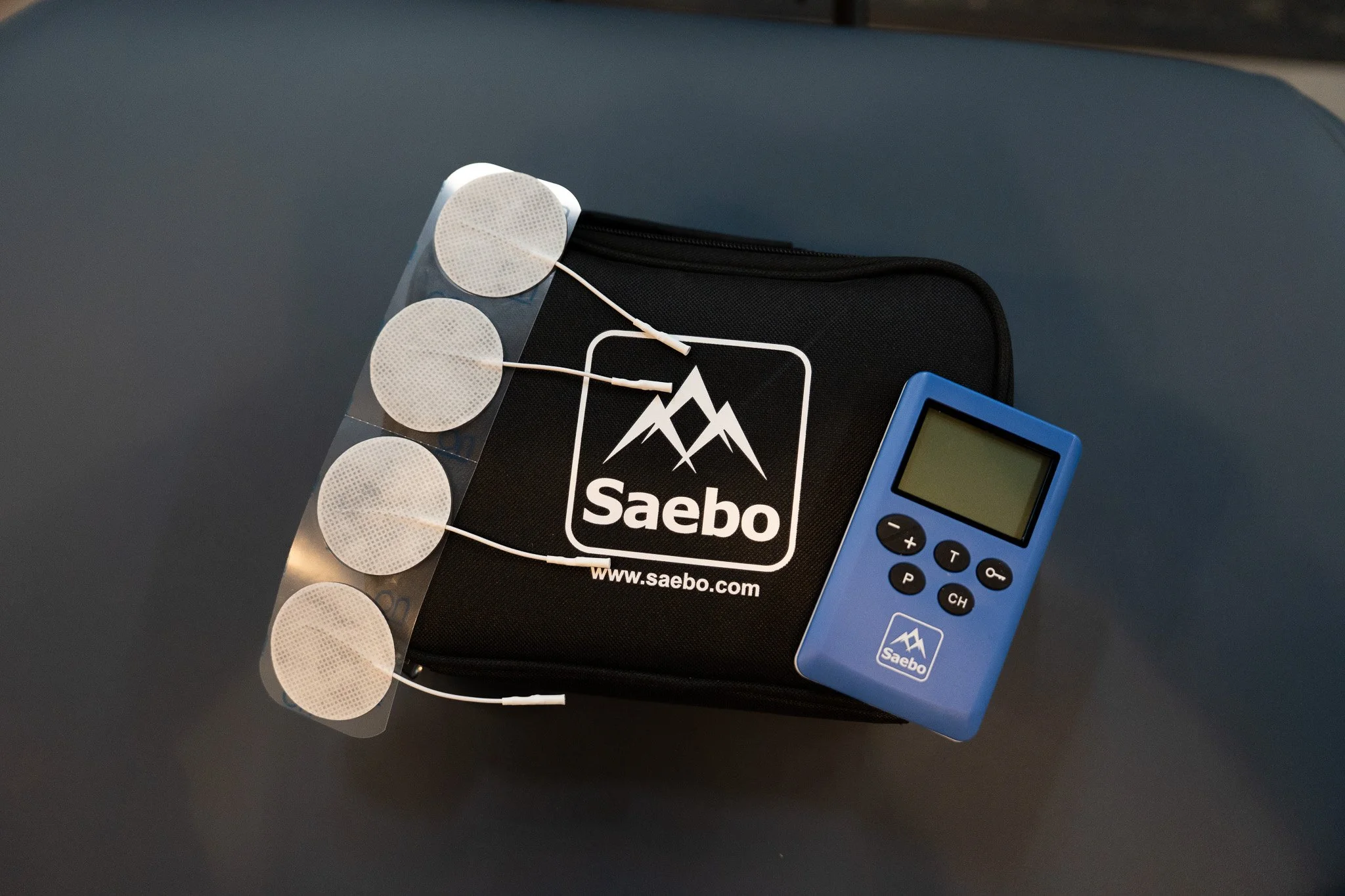 A black Saebo carrying case with a white logo, a strip of circular patches, a blue digital device with a small screen, buttons, and the Saebo logo, all placed on a dark surface.