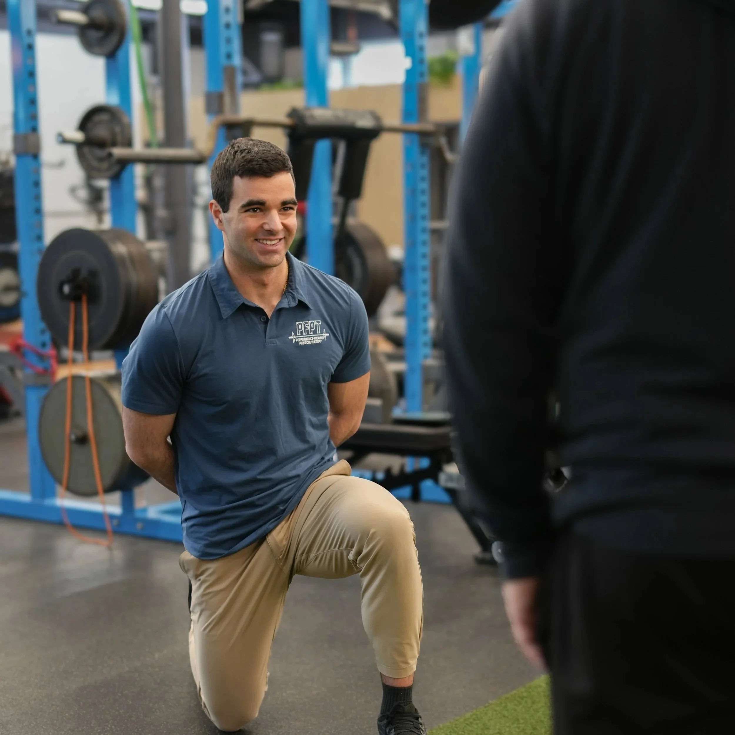 Man in blue polo shirt kneeling on one knee during a training session in a gym, with another person standing in front of him.