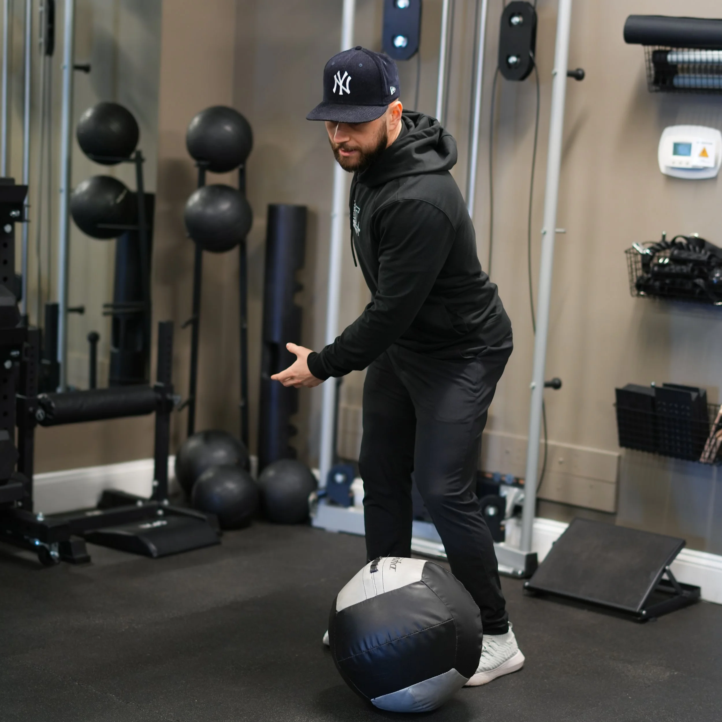 Man with beard wearing a black hoodie and a New York Yankees cap working out with a black and white medicine ball on a black gym floor in a fitness center.
