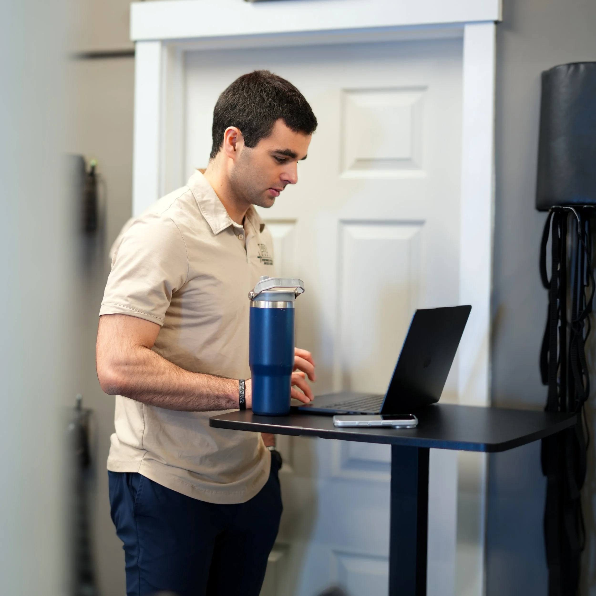 A young man with short dark hair, wearing a beige shirt and navy pants, is standing at a table working on a laptop. He has a silver and blue water bottle in front of him, and the background shows a white door and a gray wall with some mounted items.