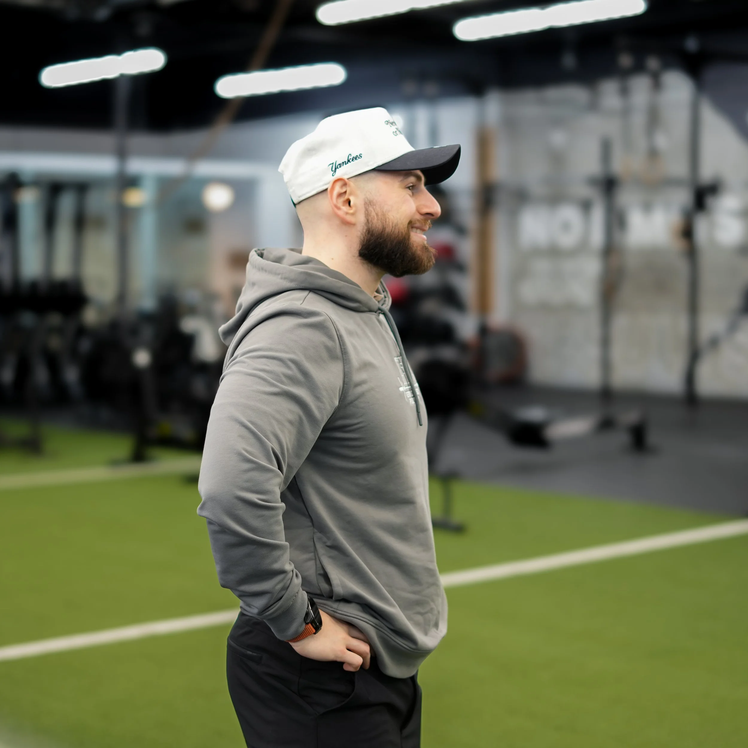 A man with a beard, wearing a gray hoodie and a white cap with black brim, standing in a gym with workout equipment and green turf in the background.