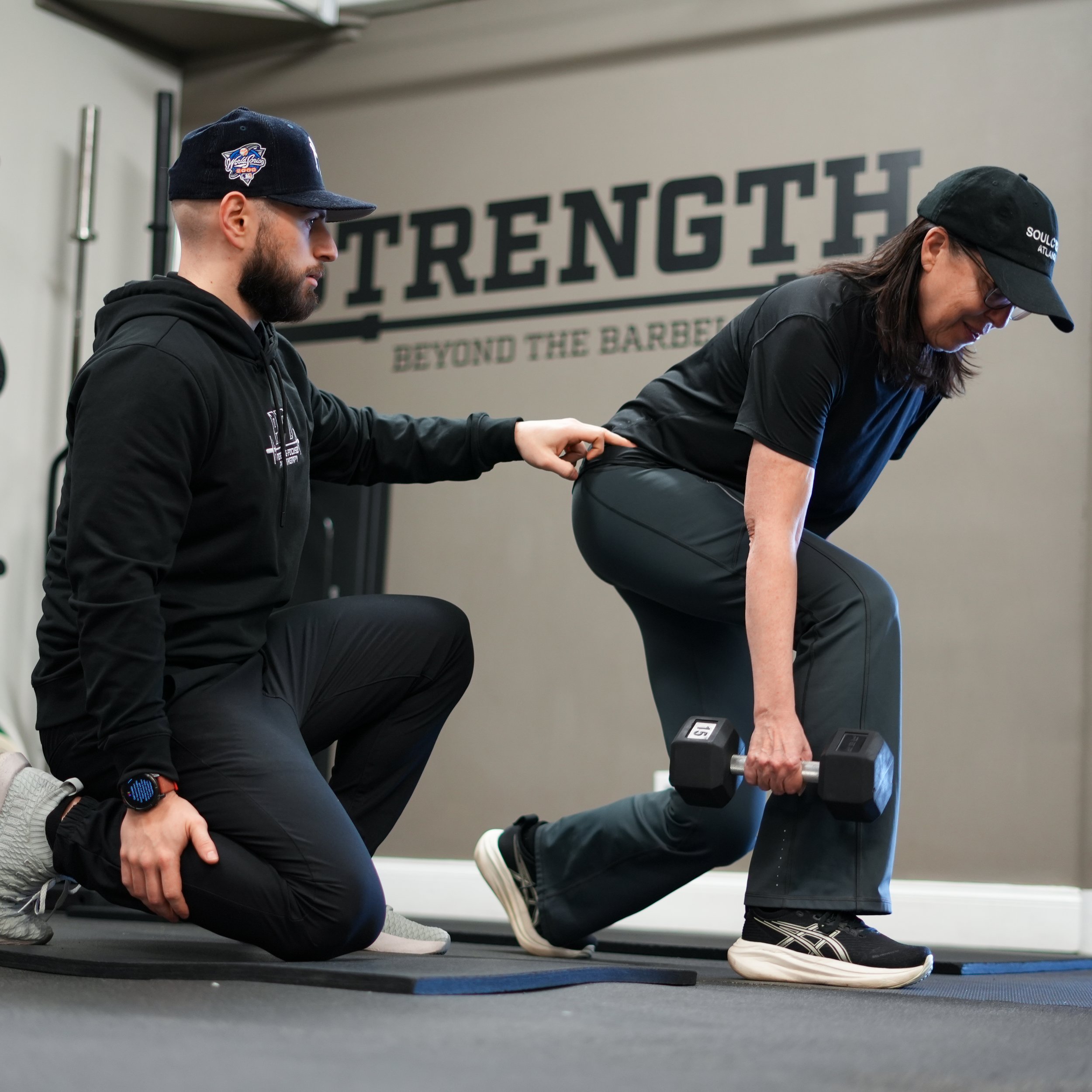A man with a beard and a woman with glasses and long hair exercising together indoors. The woman is performing a plank exercise while the man supports her by holding her arm. They are dressed in athletic clothing, and the background has a motivational sign that says 'Strength Beyond the Barriers.'