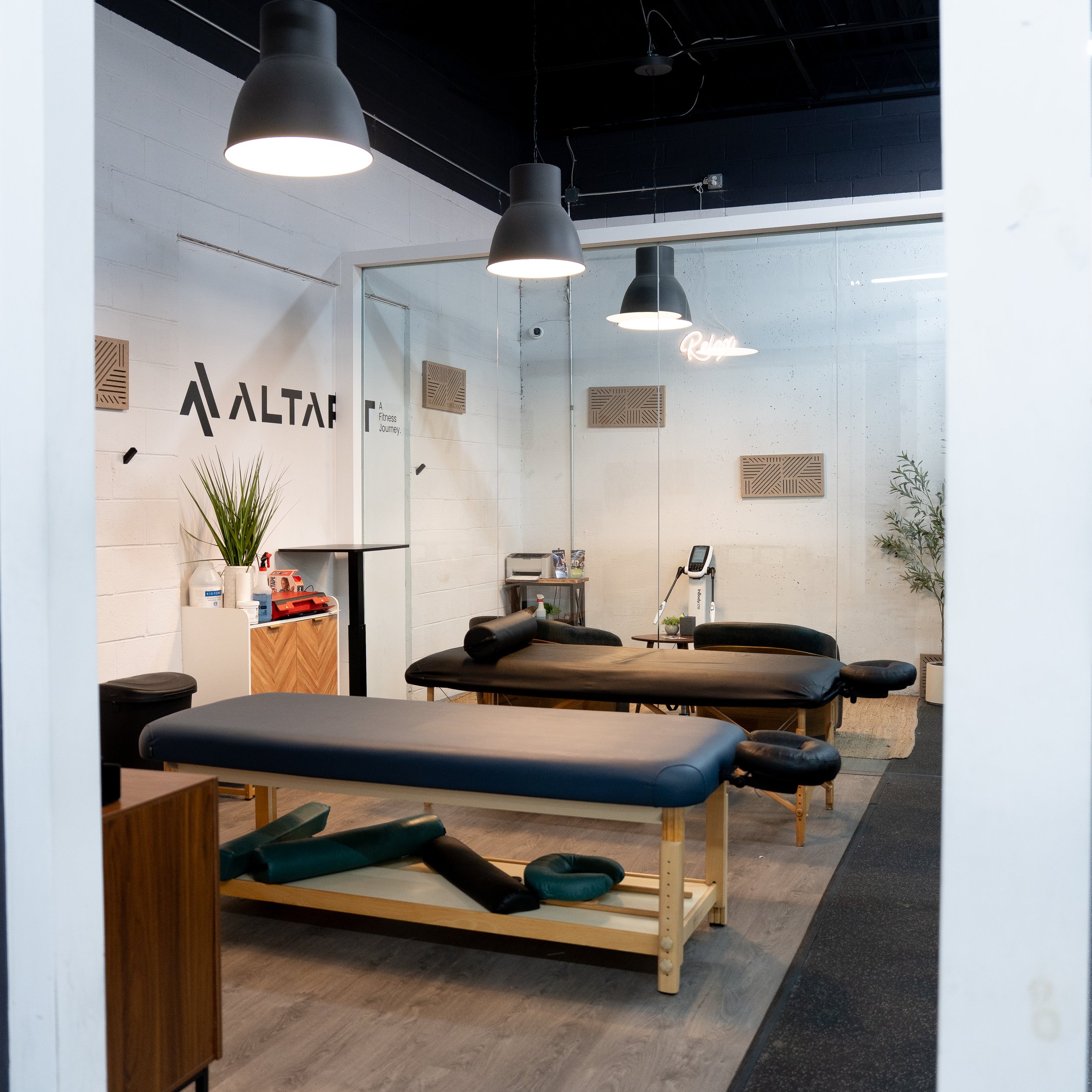 A fitness studio with exercise equipment such as mats, foam rollers, and a digital scale, illuminated by black pendant lights, featuring a mirror on the wall with the word 'Relax' written in neon, and decorated with wooden wall panels and indoor plants.
