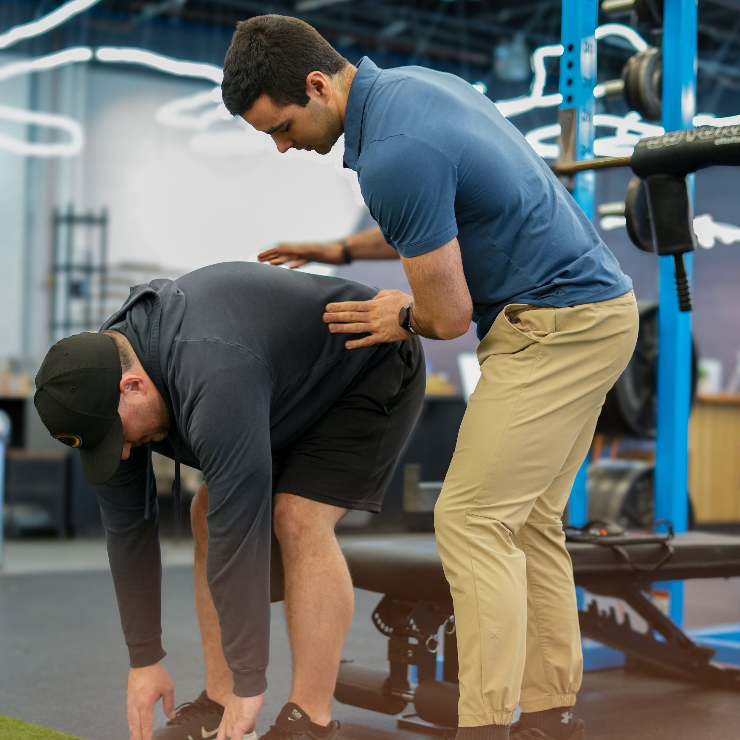 A man in a blue polo shirt and tan shorts stretching in a gym or fitness center, with a person beside him also stretching. The background shows gym equipment and blue structural supports.