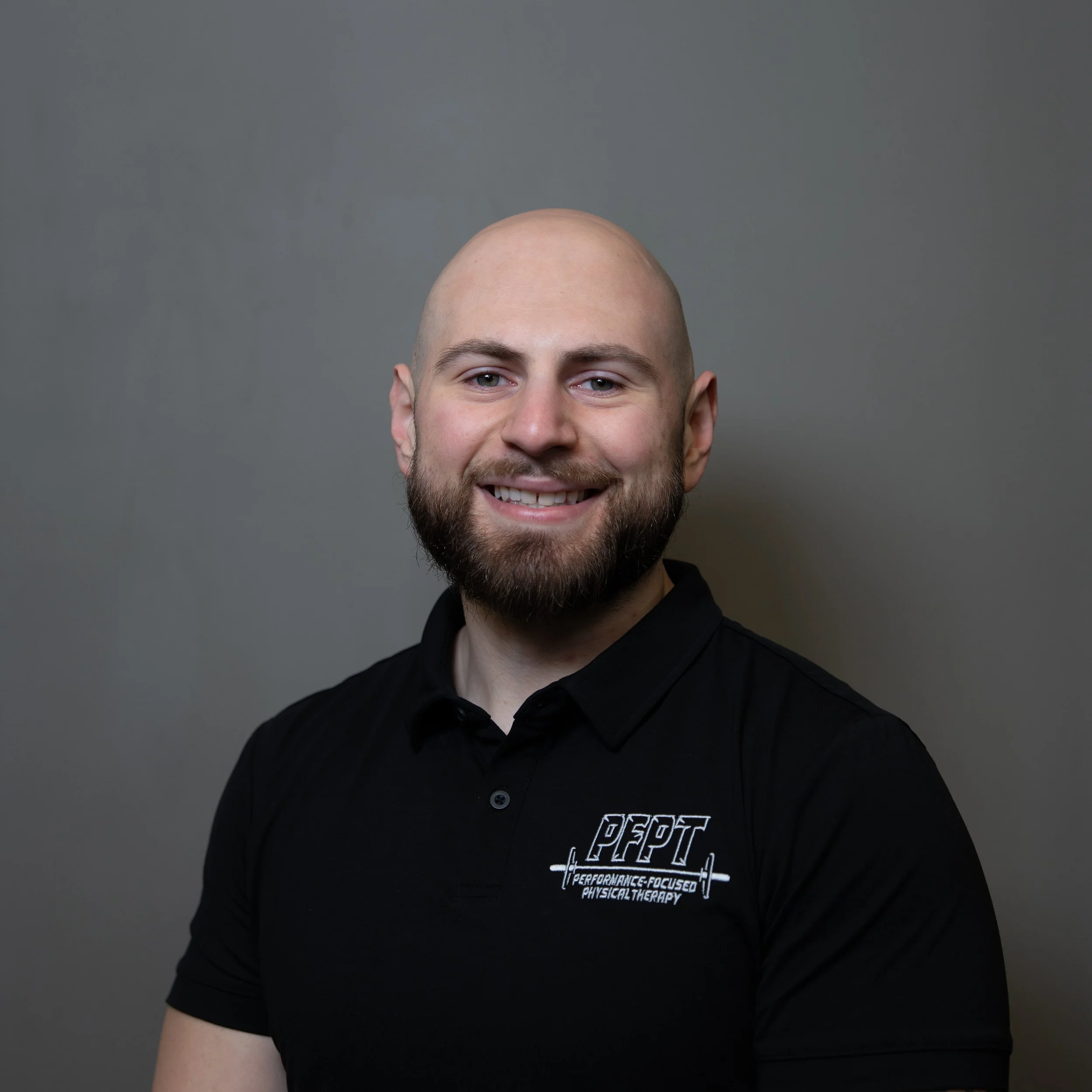 Portrait of a smiling man with a beard, wearing a black polo shirt with 'PFPT Performance Focused Physical Therapy' logo, against a plain gray background.