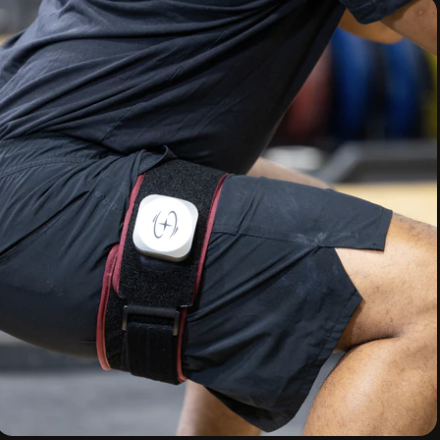 A person wearing black workout shorts with a white device attached to a black and red strap around their thigh, likely used for fitness tracking or health monitoring.