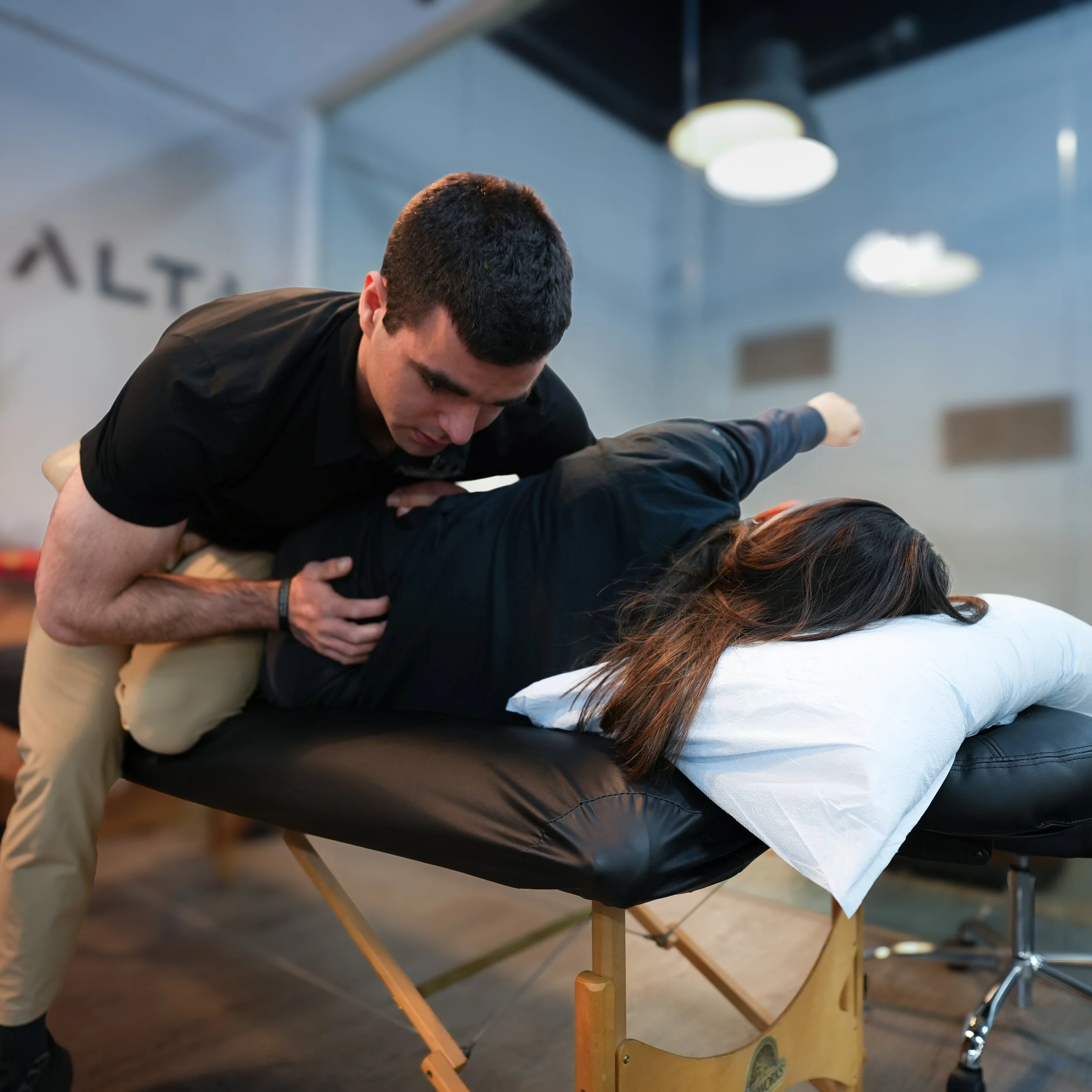 A person performing a backbending yoga pose on a massage table with their legs elevated and supported by another person's hands, in a modern indoor setting with soft lighting and a blurred background.