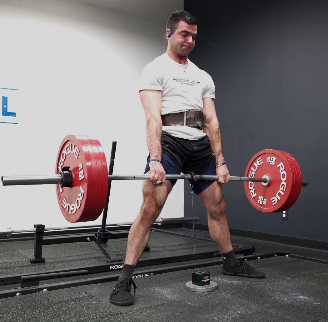 Man wearing a weightlifting belt is performing a deadlift exercise in a gym, with red weight plates on a barbell.