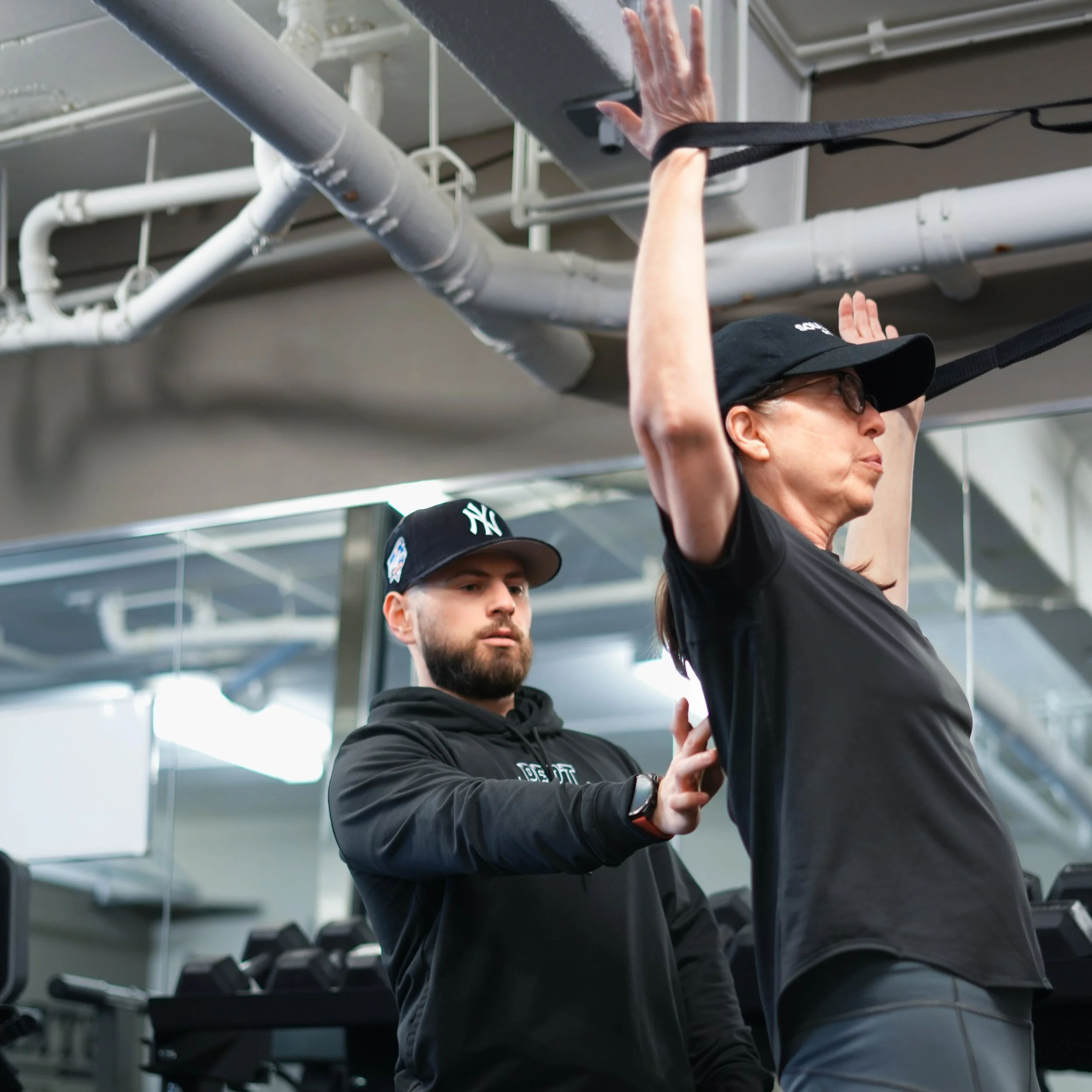 A woman is hanging from a pull-up bar in a gym, with her arms extended upward and wearing a black cap. A man next to her is observing or assisting, with gym equipment and mirrors visible in the background.