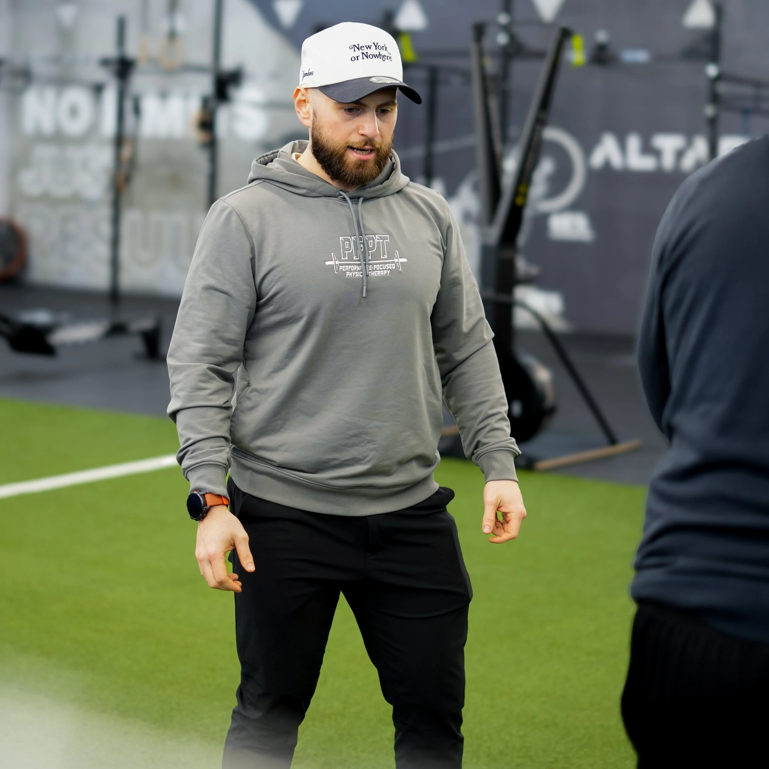 A man with a beard in athletic clothing, wearing a white and black cap and a gray hoodie, standing indoors on a green turf near gym equipment.