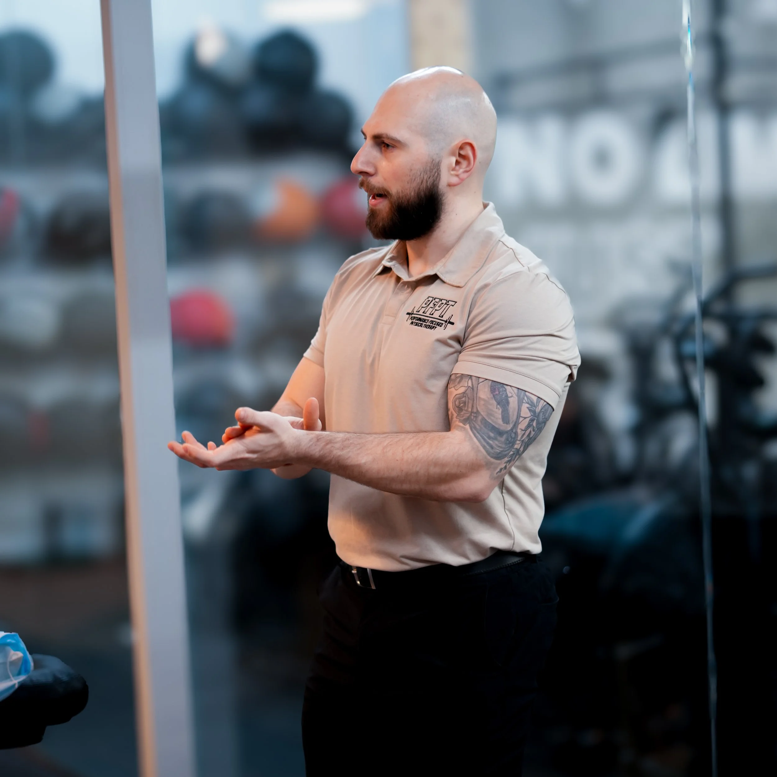 A man with a shaved head and a beard, wearing a beige shirt with logo and slogan, is standing in a gym with blurred fitness equipment in the background, possibly engaged in a conversation or listening attentively.