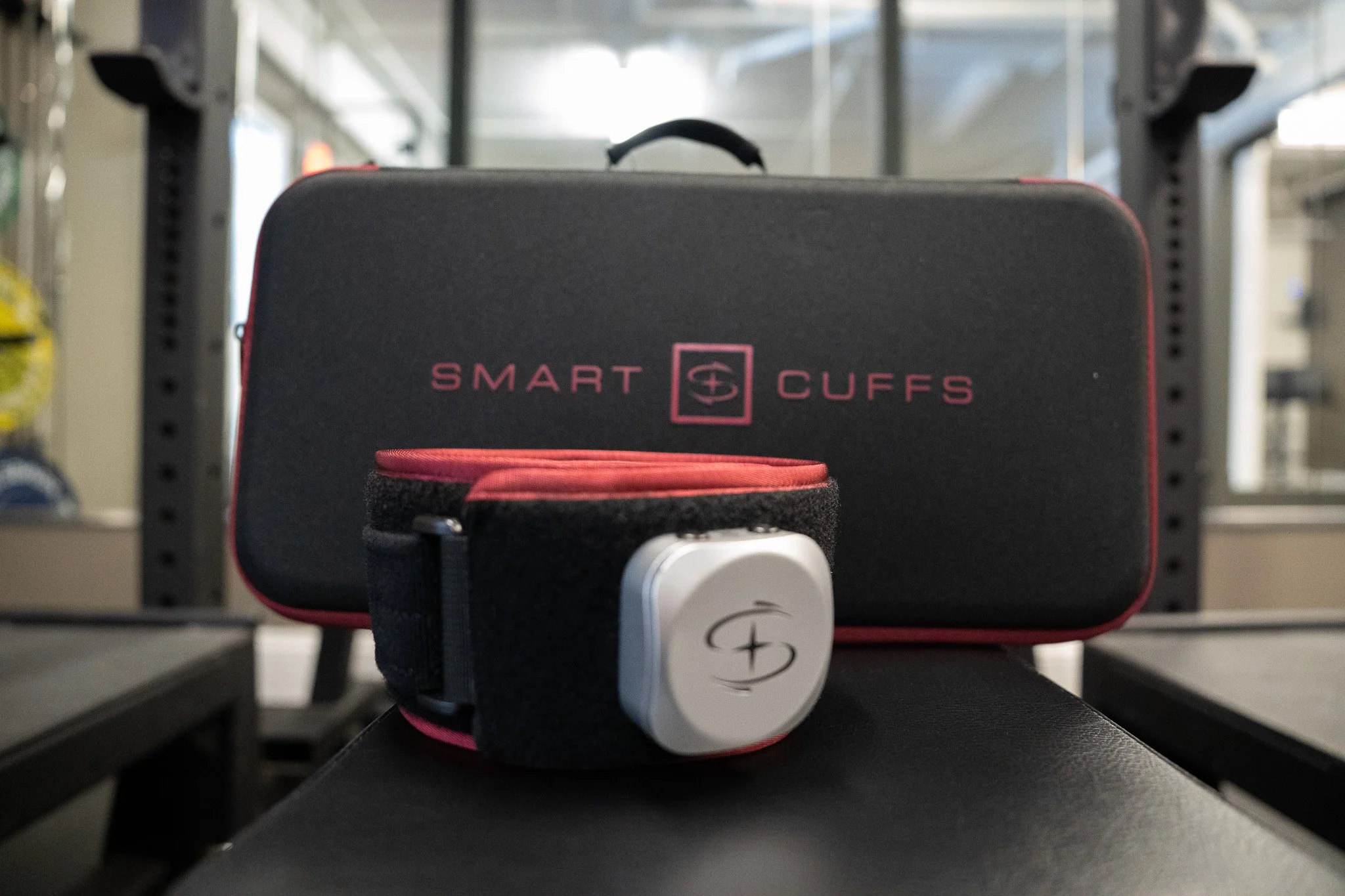 A close-up of a black protective case labeled 'Smart Cuffs' with pink text and logo, placed on a black surface with fitness equipment in the background.