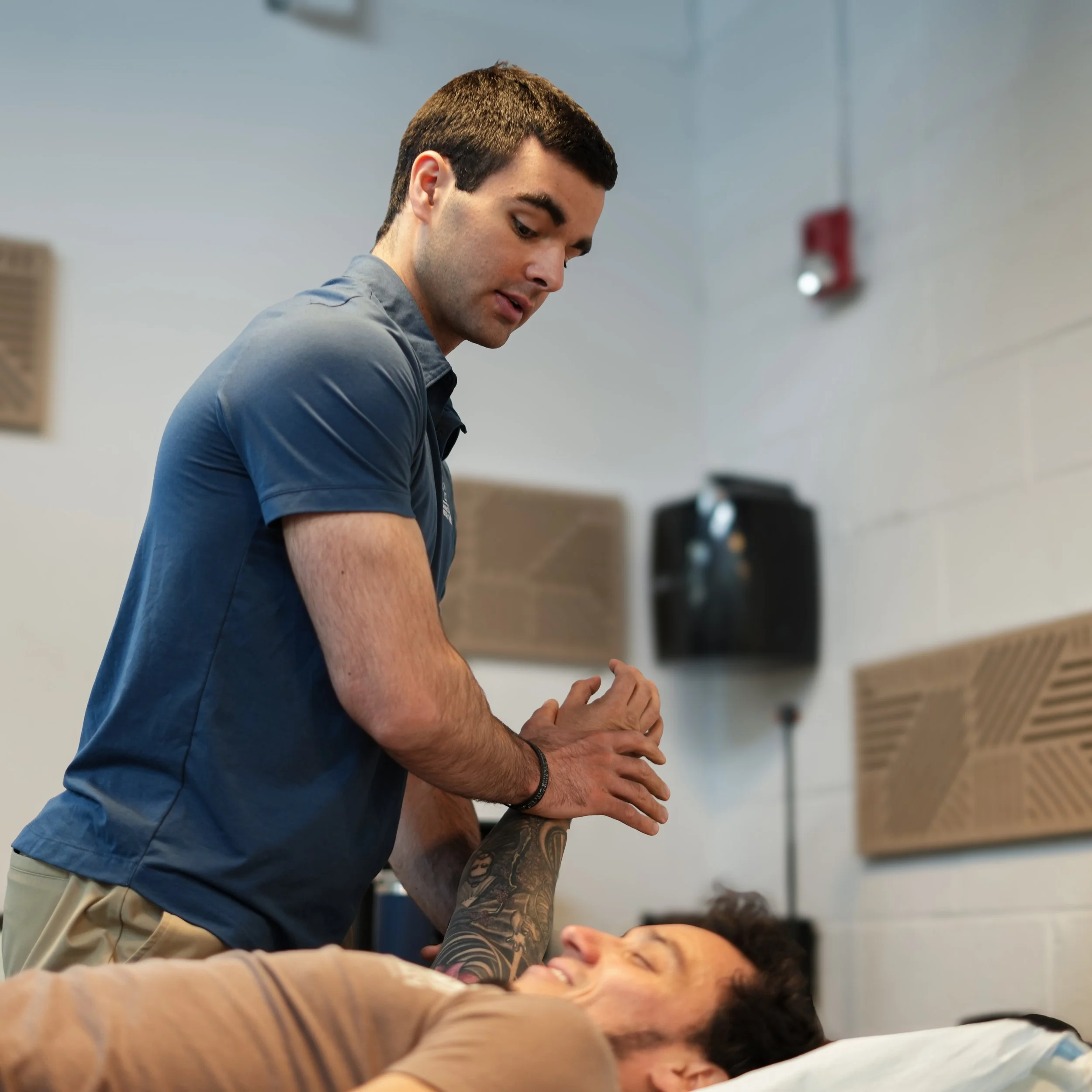 A young man with short brown hair, wearing a gray shirt, is leaning over a woman lying on a table, possibly in a medical or tattoo studio. The woman has dark hair and is smiling, appearing relaxed. The background shows a plain wall with a red fire alarm or sprinkler system.
