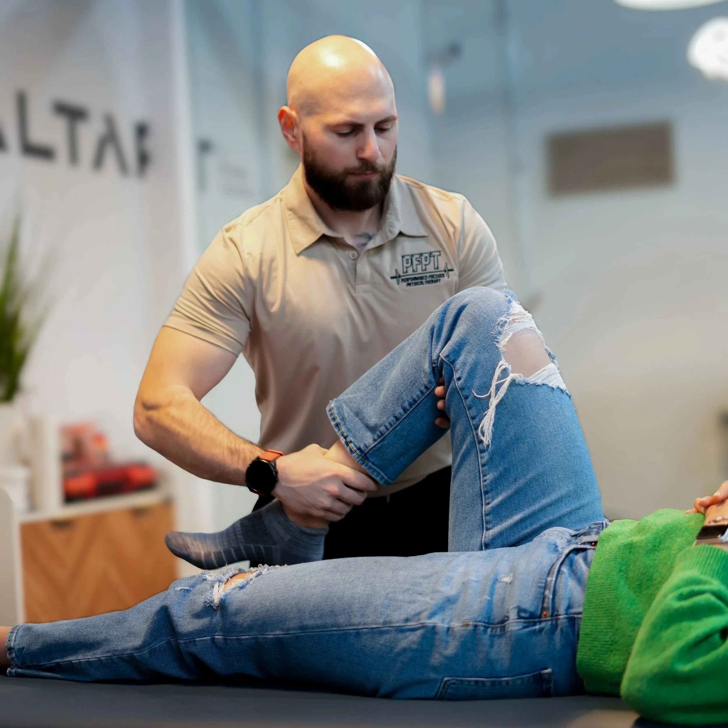 A man with a shaved head and beard wearing a beige polo shirt with a logo, sitting cross-legged on the floor, looking downward.