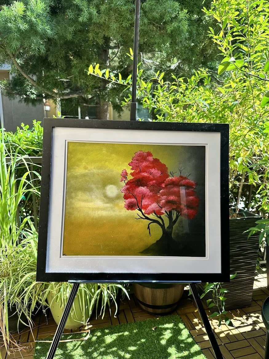 “Emerging from a muted golden mist, the red tree becomes a symbol of resilience and beauty. Its bright foliage contrasts with the calm, hazy background, creating a peaceful moment of reflection.” - painting by Neil Banerjee