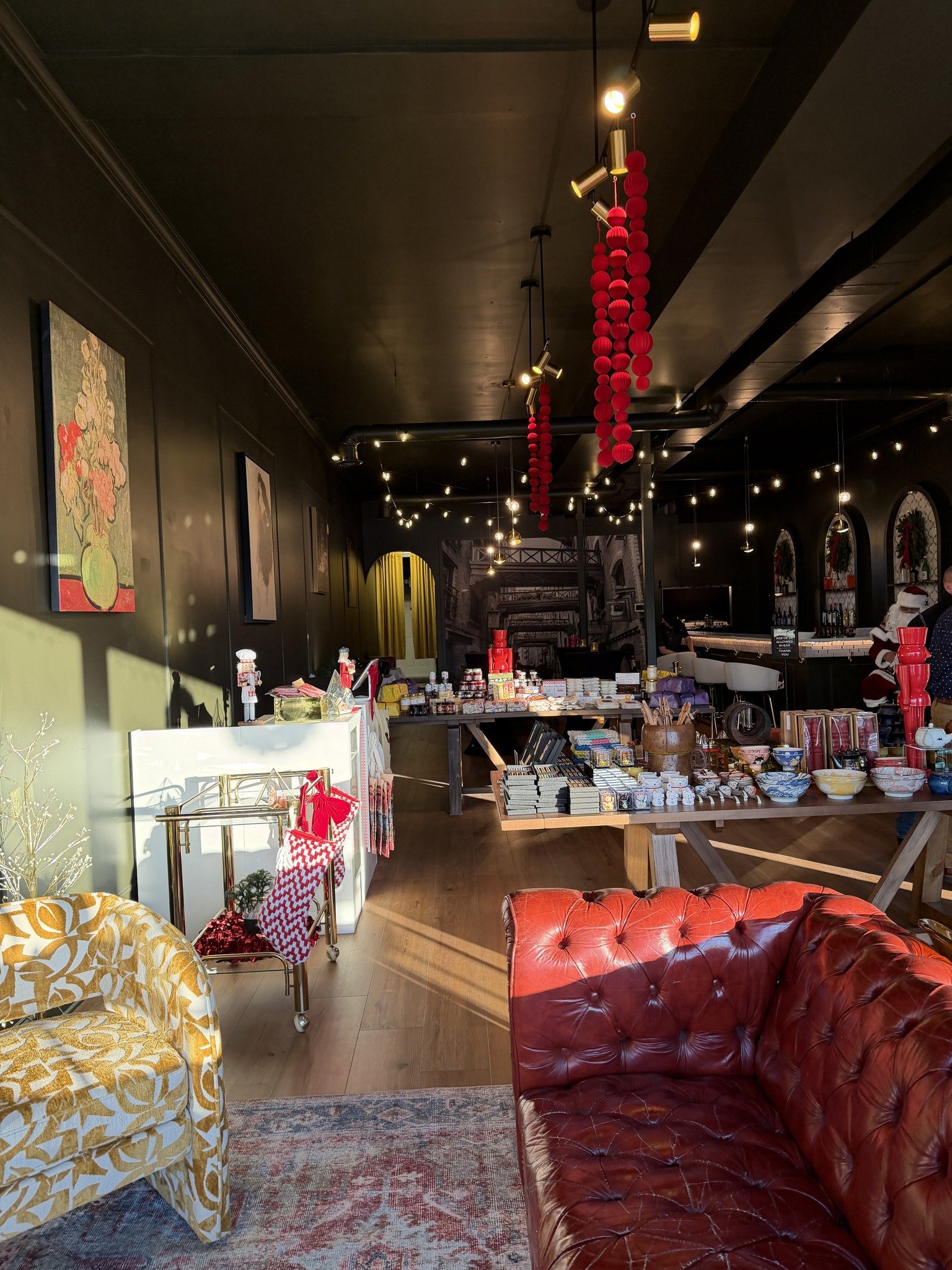 Interior of a decorated shop with Christmas decorations, including red ornaments hanging from the ceiling, and tables displaying various gift items. There are cozy chairs and patterned sofas in the foreground, and a bar area with shelves in the backg