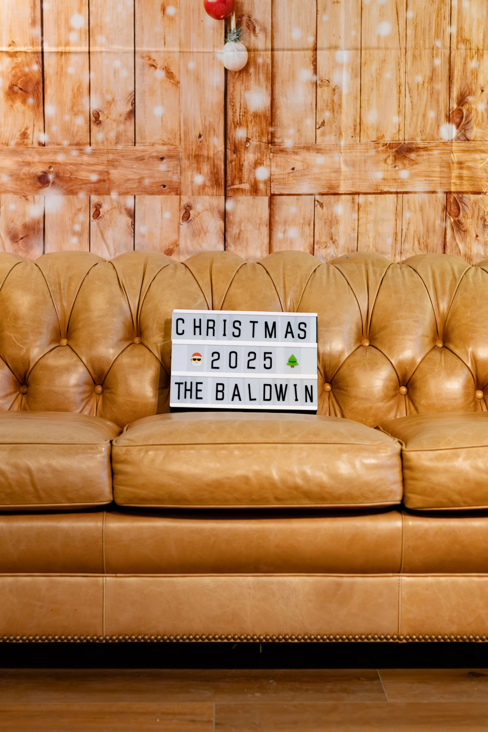 A tan leather tufted sofa against a wooden wall. On the sofa, there is a lightbox with black letters spelling out "CHRISTMAS 2025 THE BALDWIN," with Christmas-themed emojis of sunglasses and a tree.