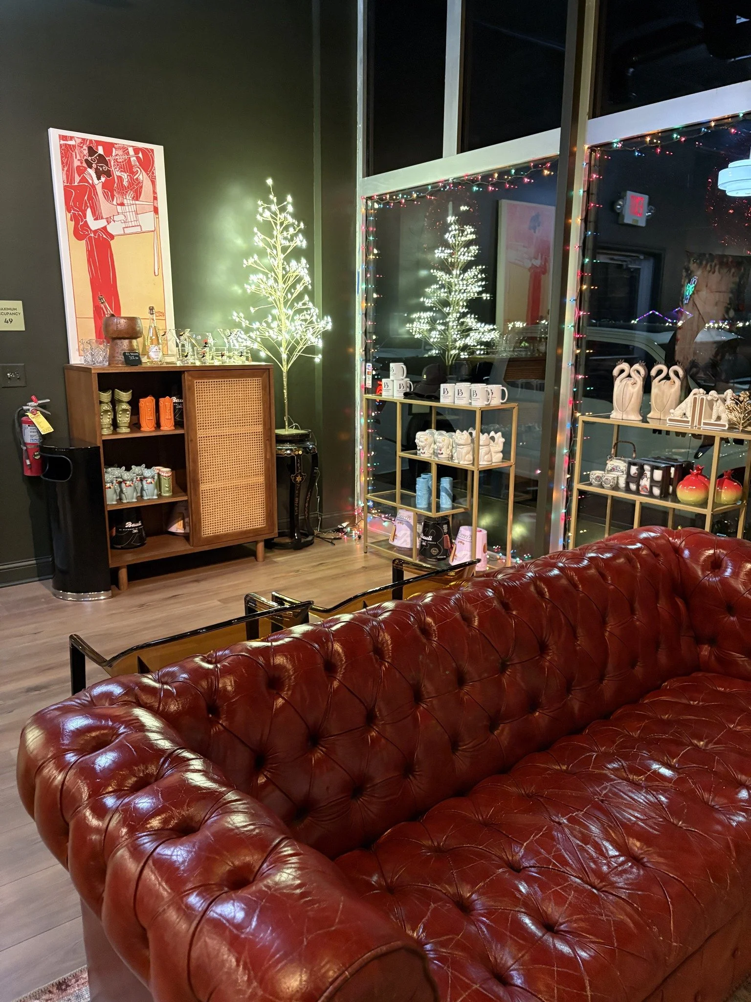 Interior of a cozy shop or cafe, decorated with string lights and two illuminated artificial Christmas trees. A red leather tufted sofa is in the foreground, and shelves with various decorative items and mugs are visible in the background.