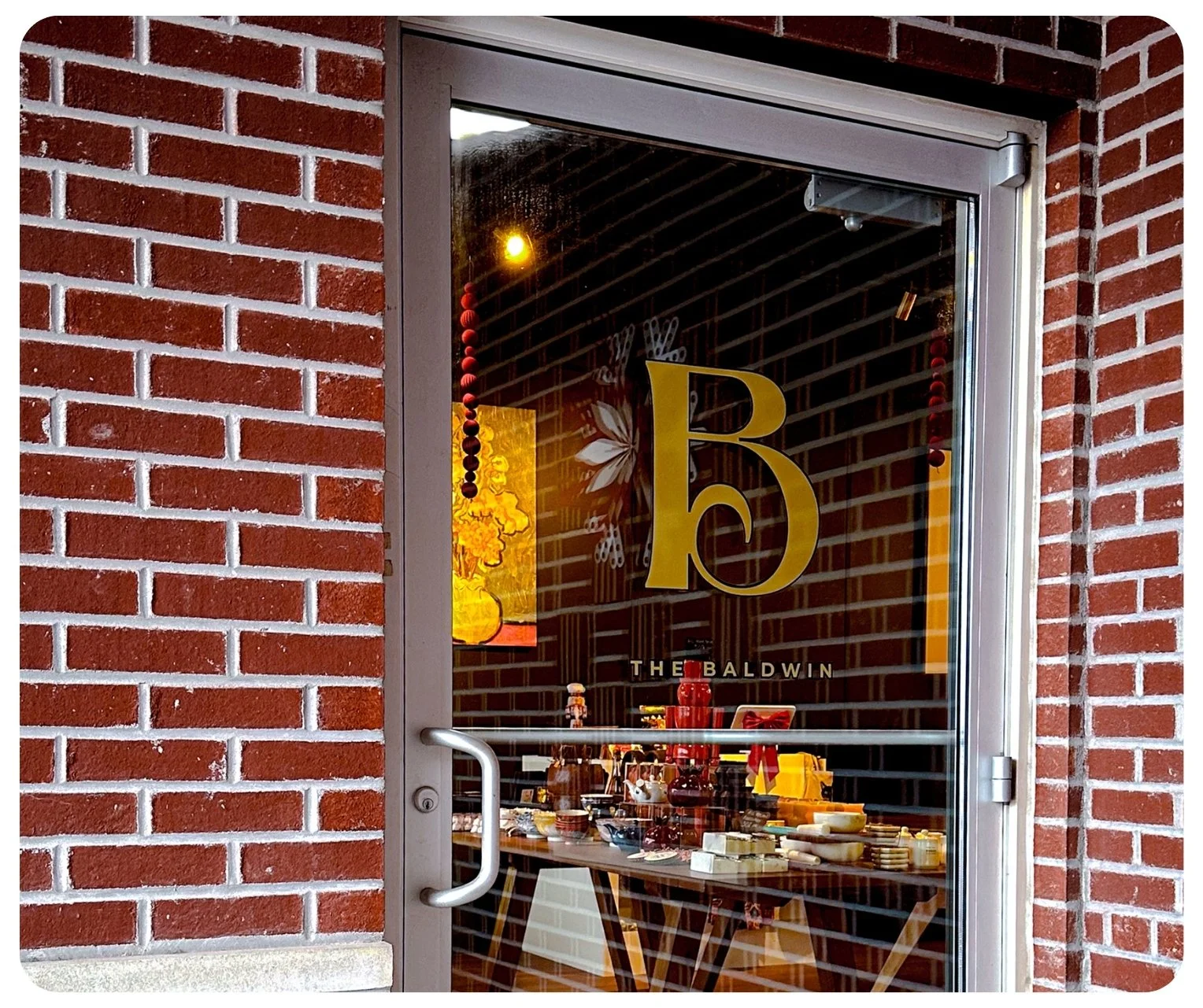 Restaurant door with glass window showing a yellow logo with the letter 'B' and the words 'The Baldwin' underneath. Inside, there are tables with various dishes and decorations surrounding a hanging yellow lamp.