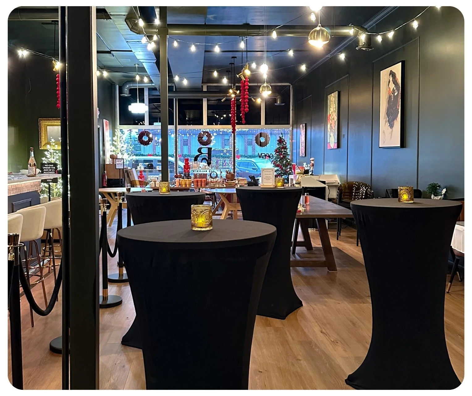 Interior of a restaurant decorated for Christmas with black high-top tables, candles, and a small Christmas tree. Front window has festive wreaths and red garlands, with holiday decorations and string lights hanging from the ceiling.