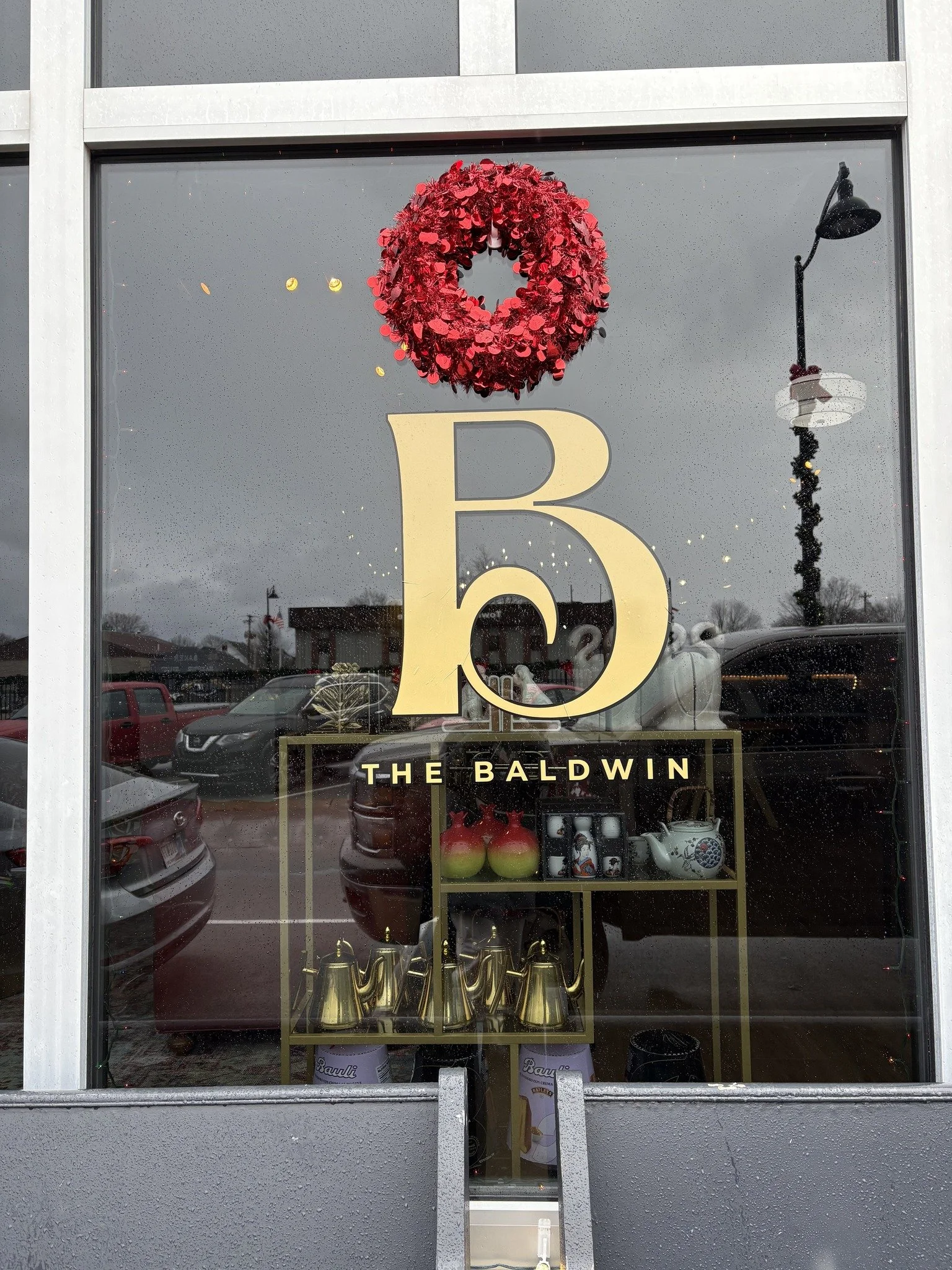 Store window with a holiday wreath, large gold letter B, and text 'The Baldwin'; inside are decorative items including teapots, apples, and gold coffee pots.
