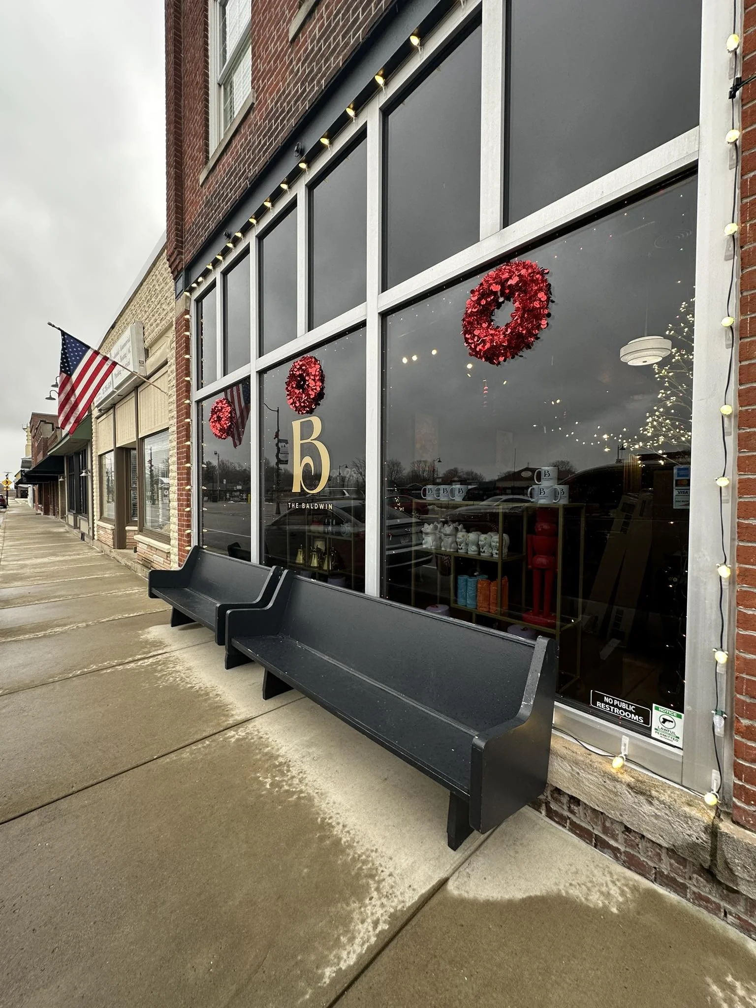 Empty black benches outside a storefront decorated with red wreaths and string lights, American flags hanging on the building, and a large letter 'B' on the window.