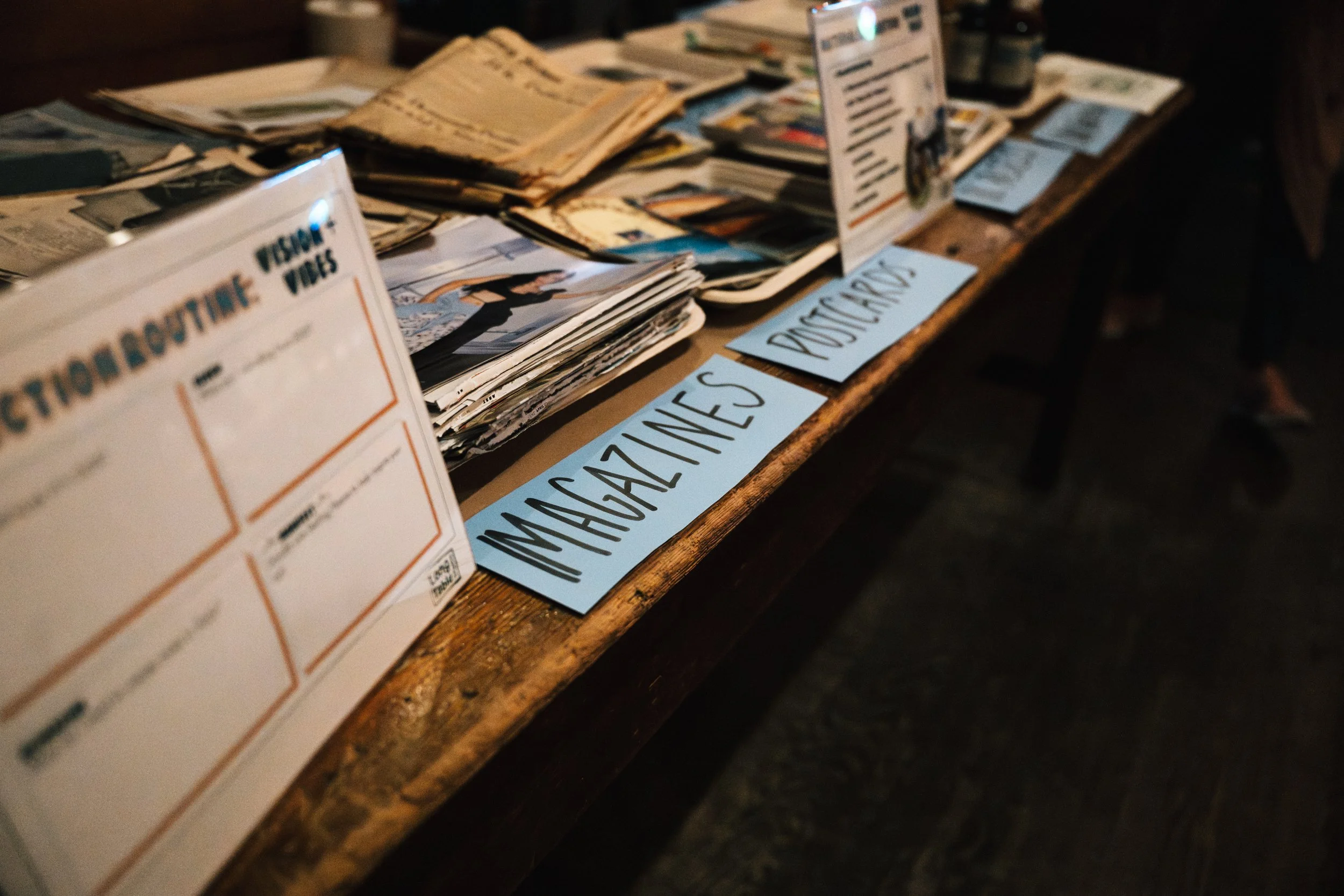 A wooden table with stacks of newspapers and magazines, and handwritten signs reading 'MAGAZINES' and 'POSTCARDS.'